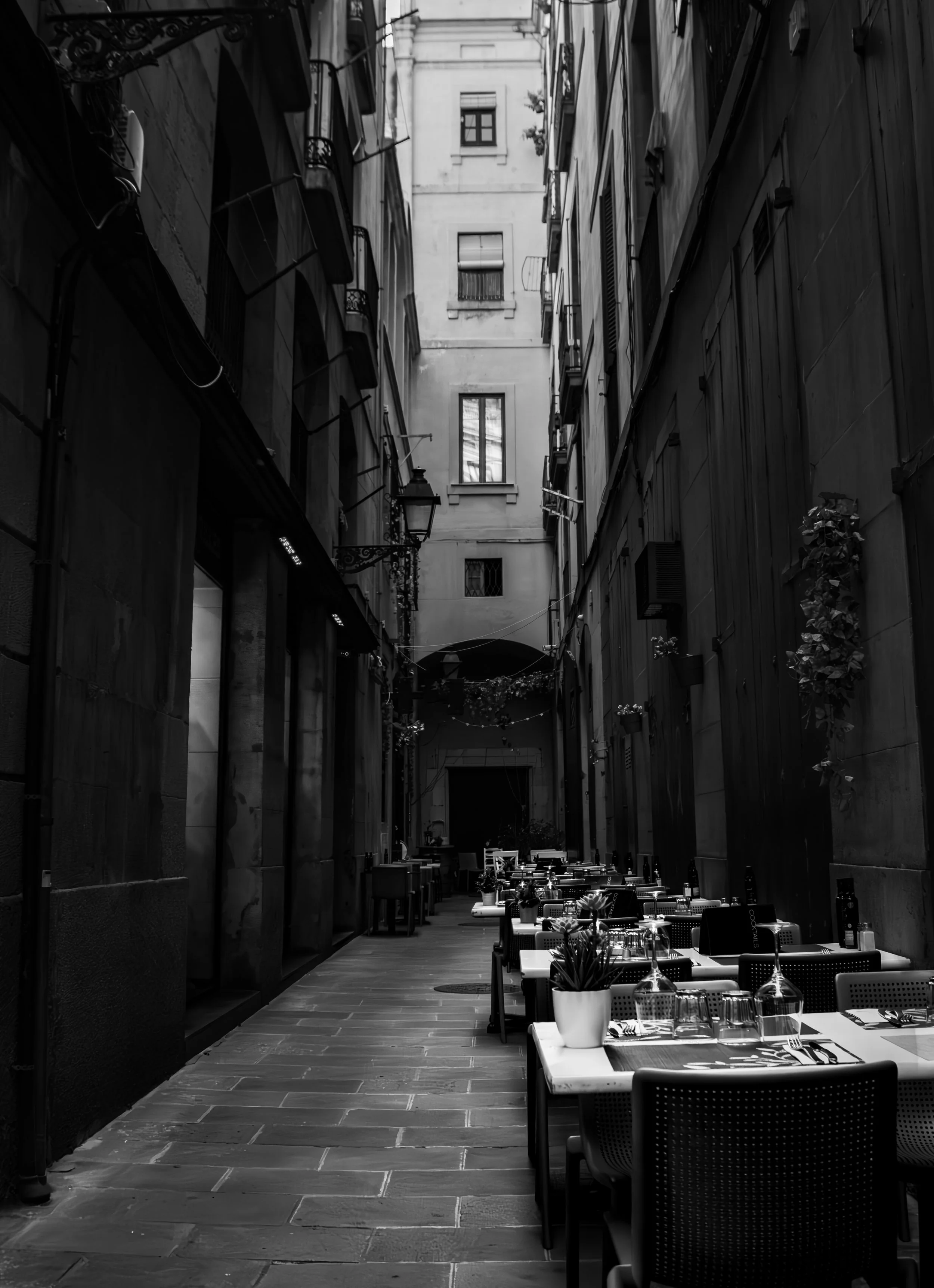 Outdoor restaurant patio with tables, chairs, and potted plants in a narrow alleyway, surrounded by tall buildings.