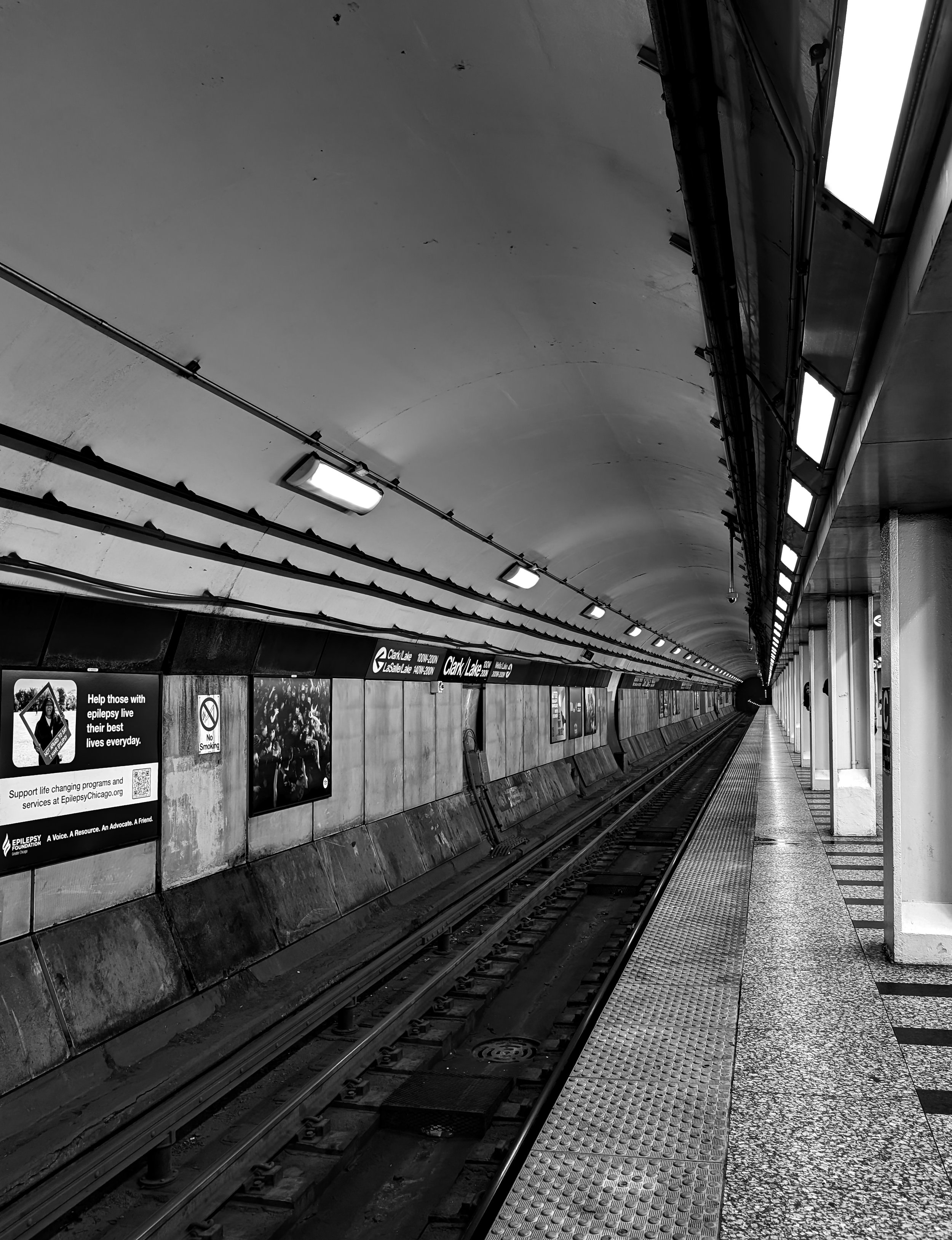 An empty subway station platform with train tracks, tiled flooring, and overhead lights, with advertisements on the wall.