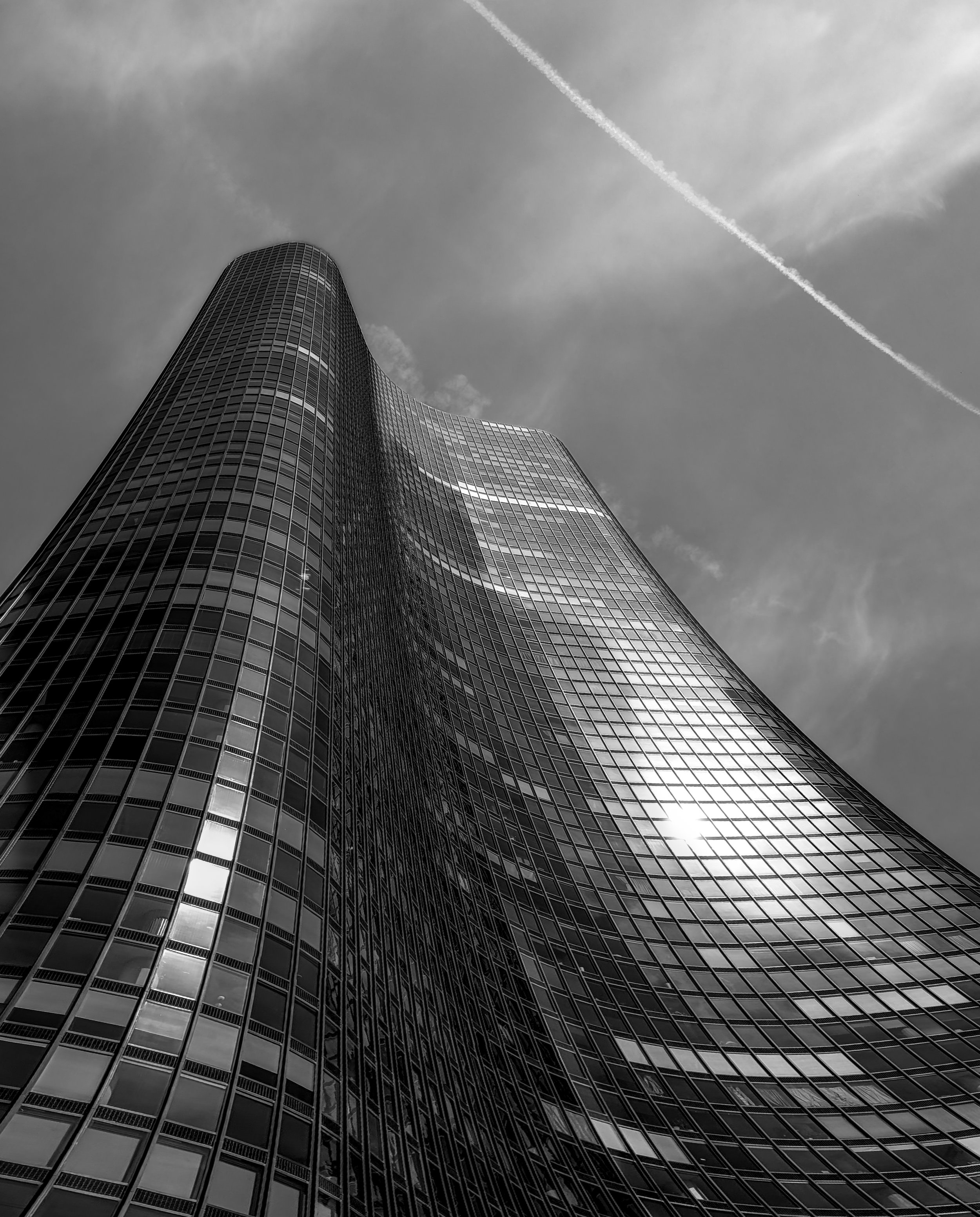 A black and white photo of a modern, curved skyscraper with reflective glass windows, taken from a low angle against a partly cloudy sky with visible airplane contrail.