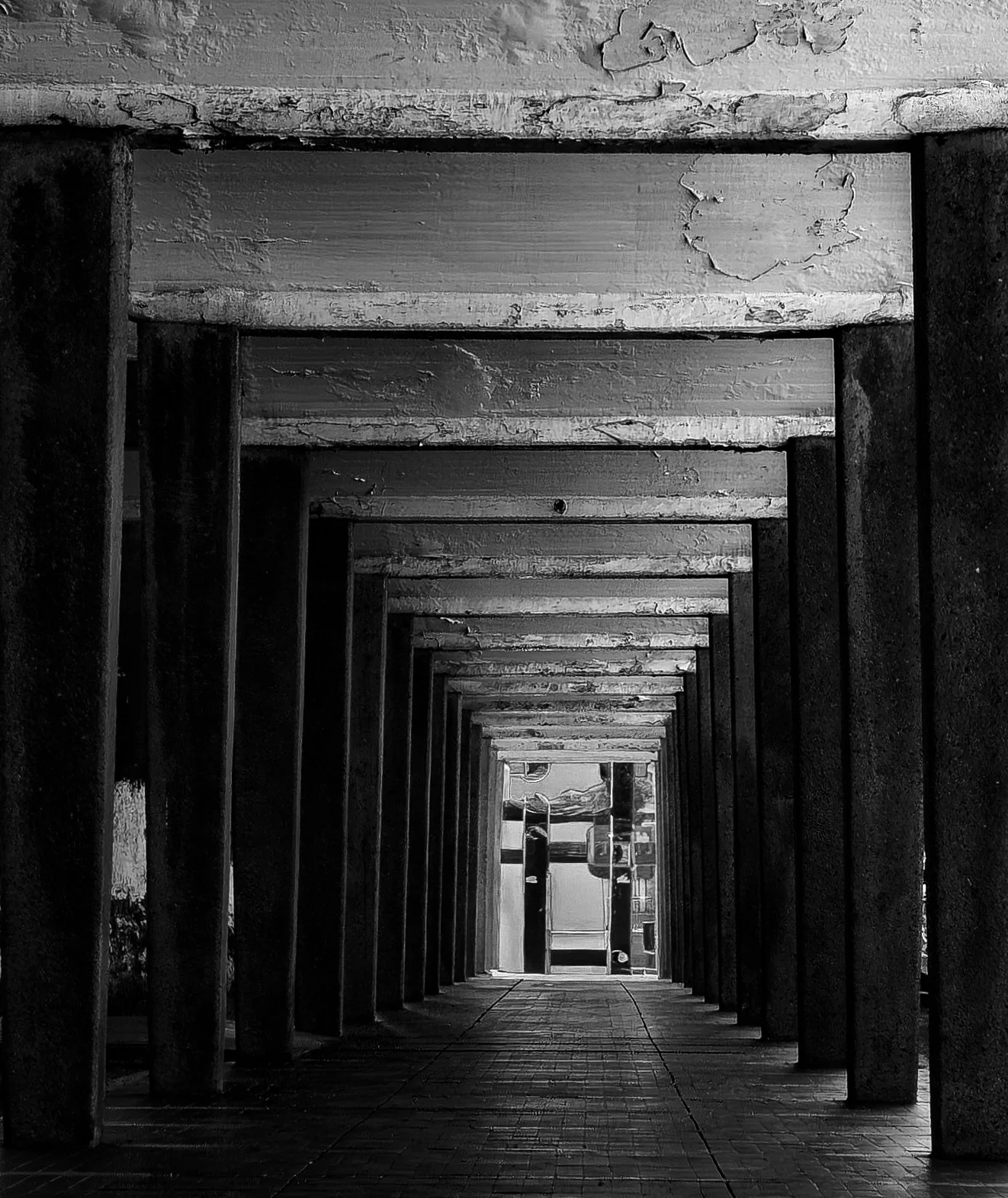 Black and white image of repeating concrete beams forming a tunnel-like structure, with strong symmetry and depth leading toward a distant opening.