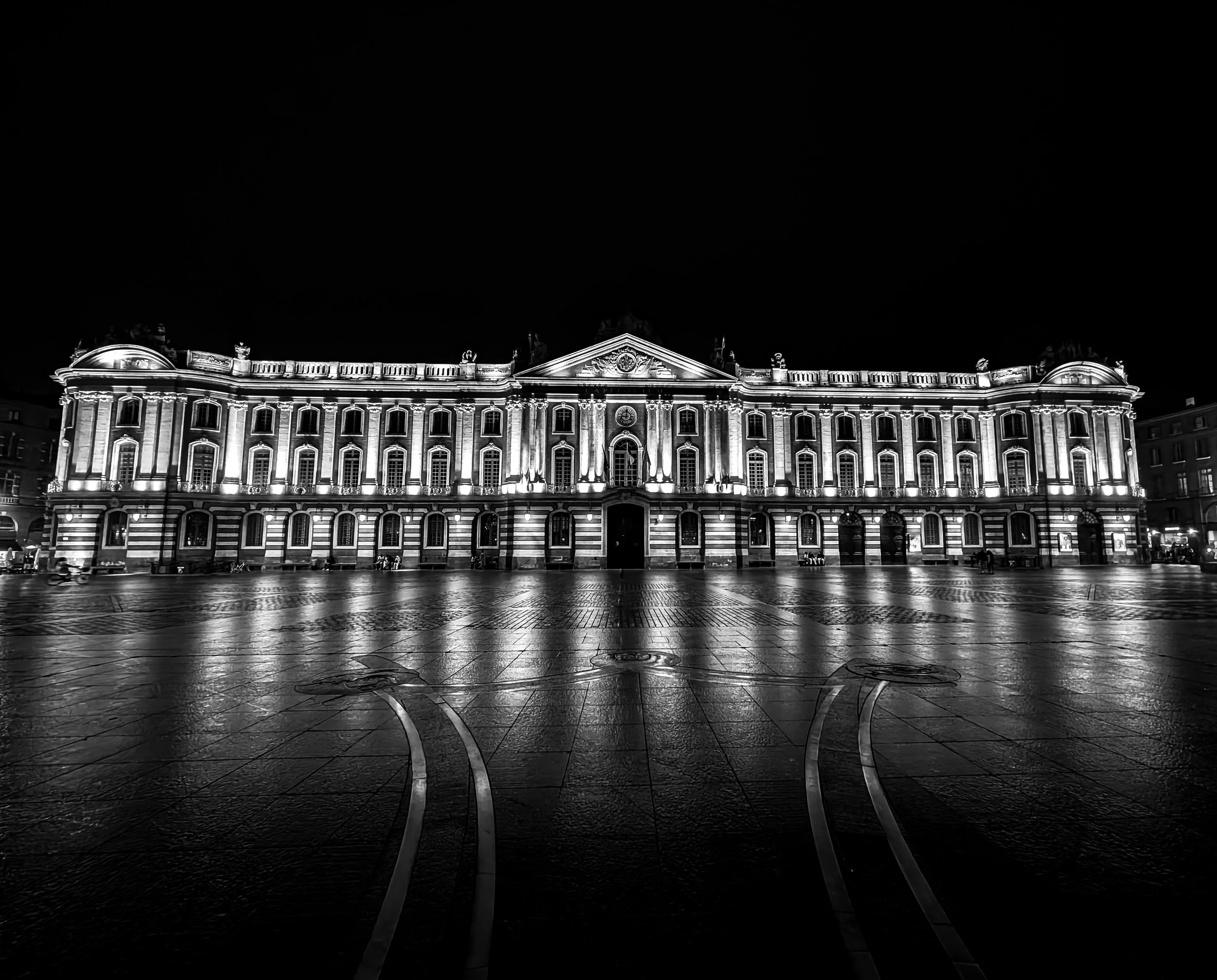 The illuminated building at night with lit windows and detailed architecture, reflecting on the wet ground in the foreground.