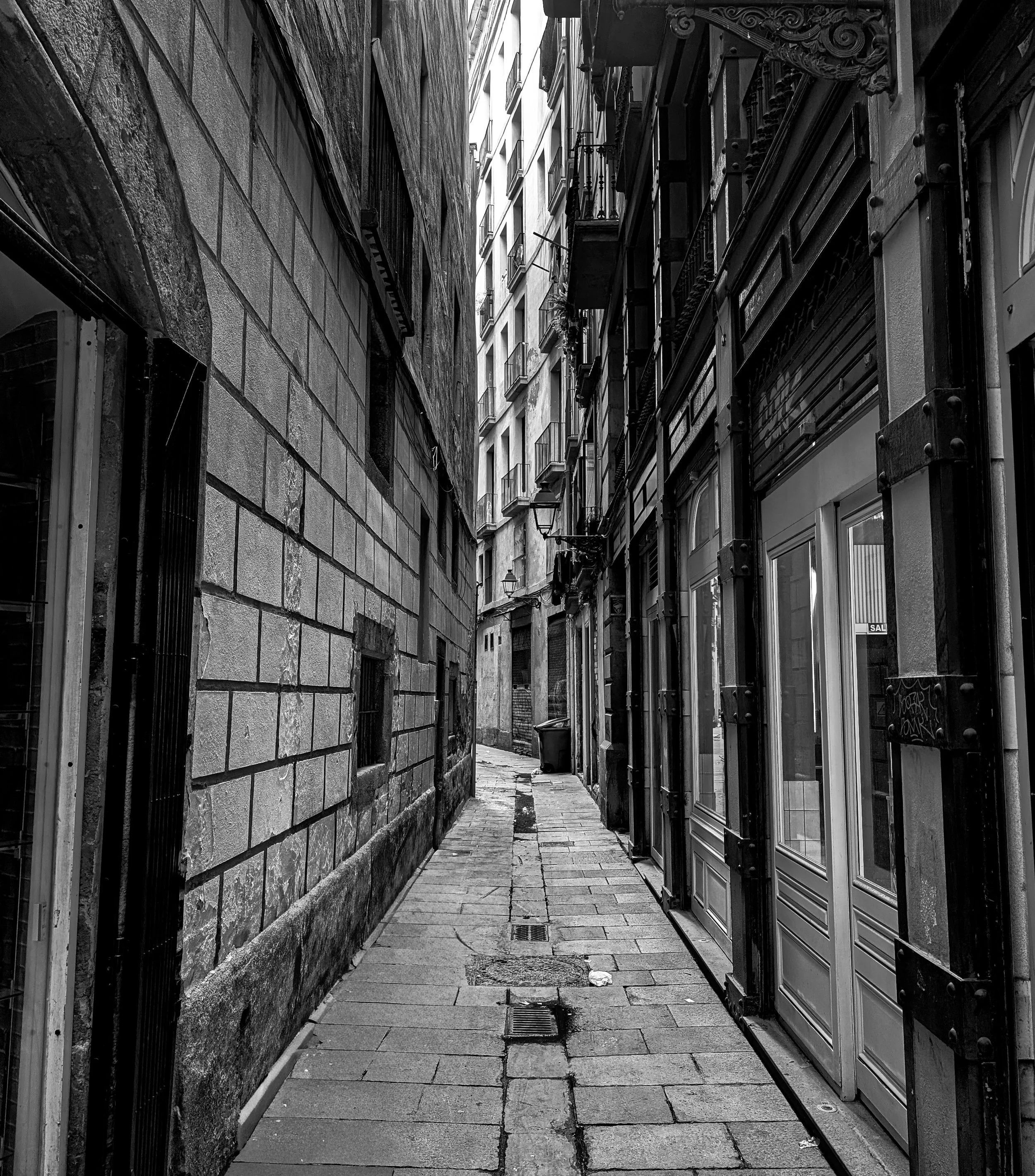 Black and white photograph of a tight urban corridor framed by tall buildings, emphasizing compressed space and vertical lines.
