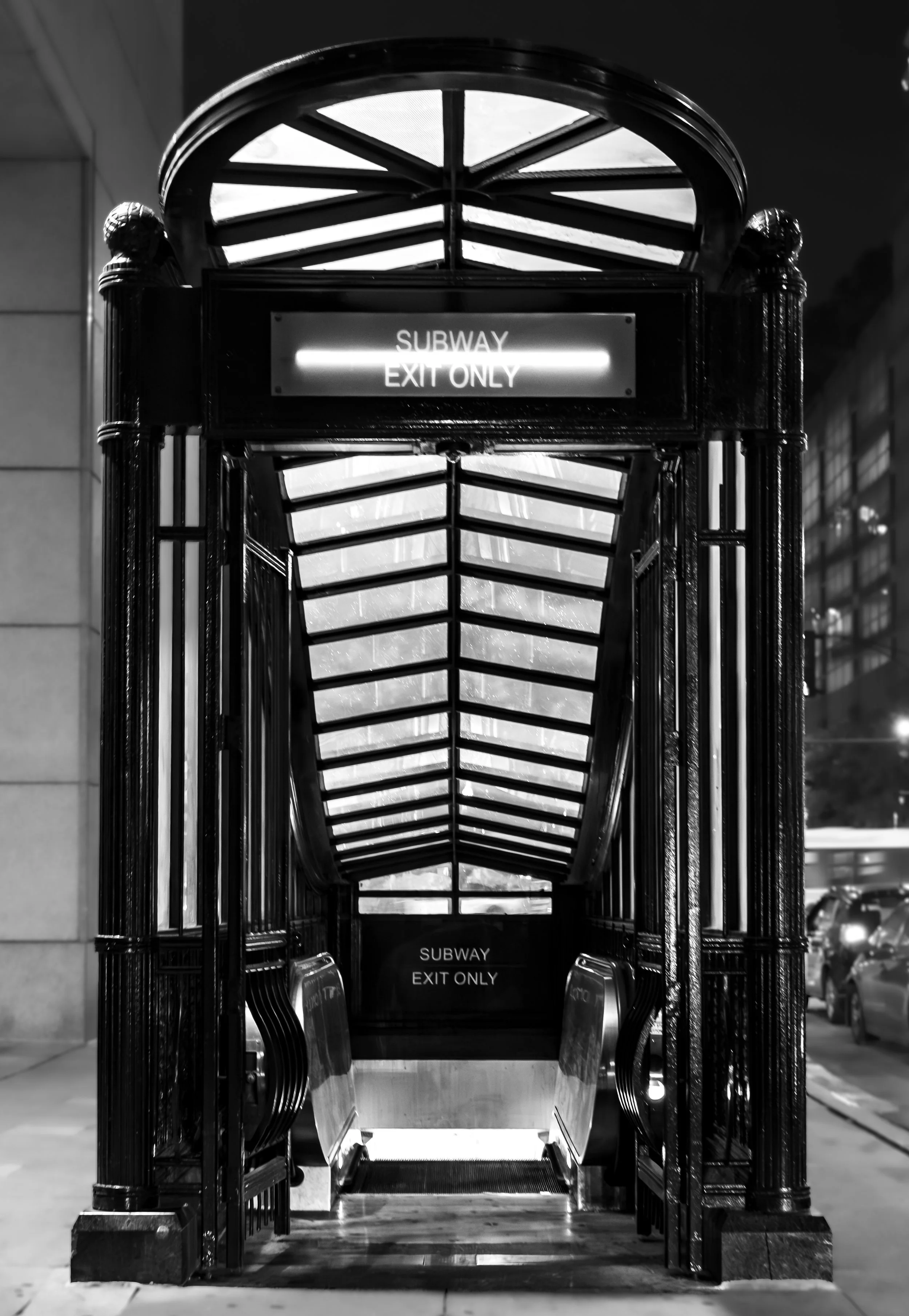 Black and white photo of an underground subway exit with a metal staircase and glass roof, featuring illuminated signs that read 'Subway Exit Only'.