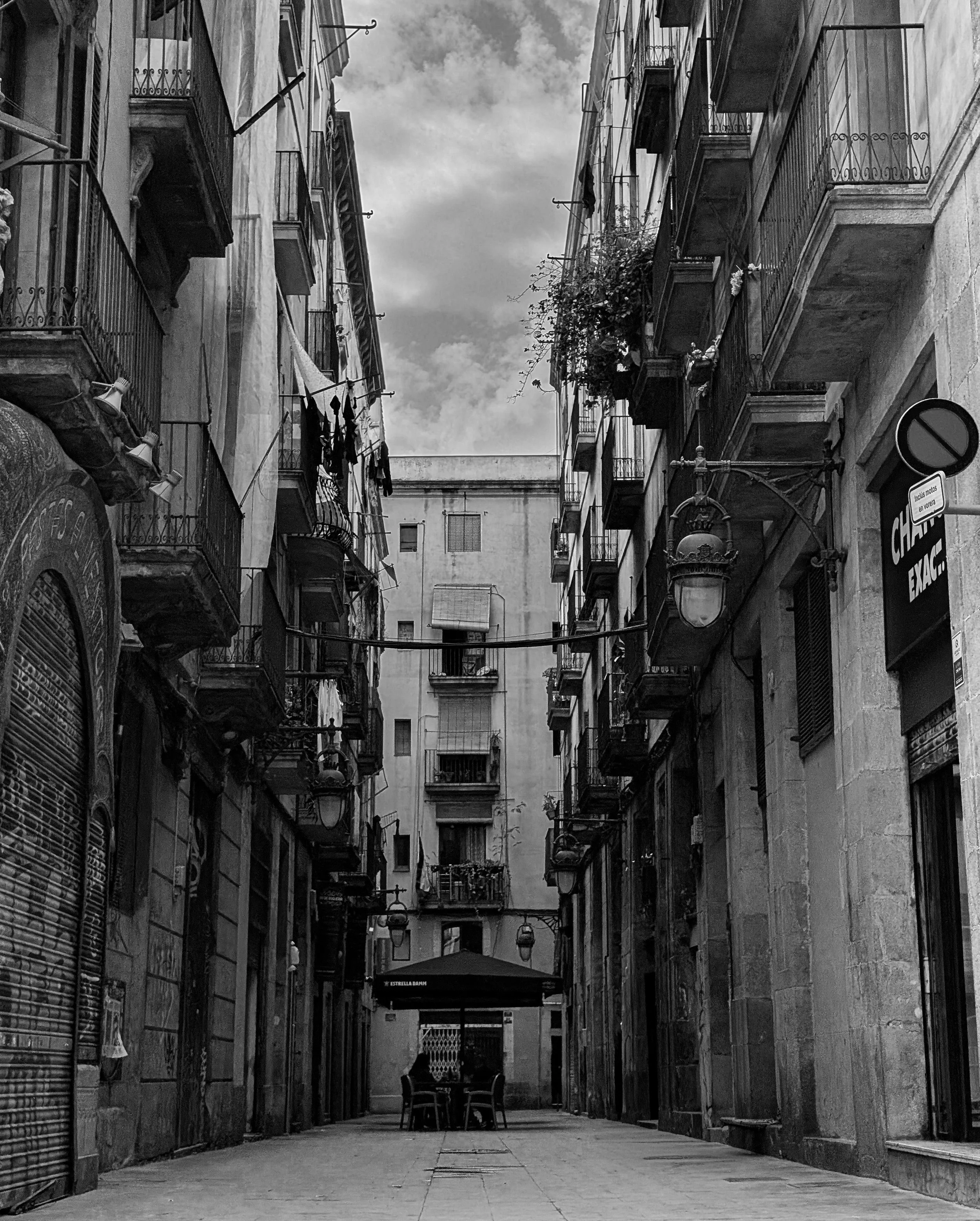 Black and white photograph of a narrow alley framed by tall buildings, leading toward a small seating area at the end of the passage.