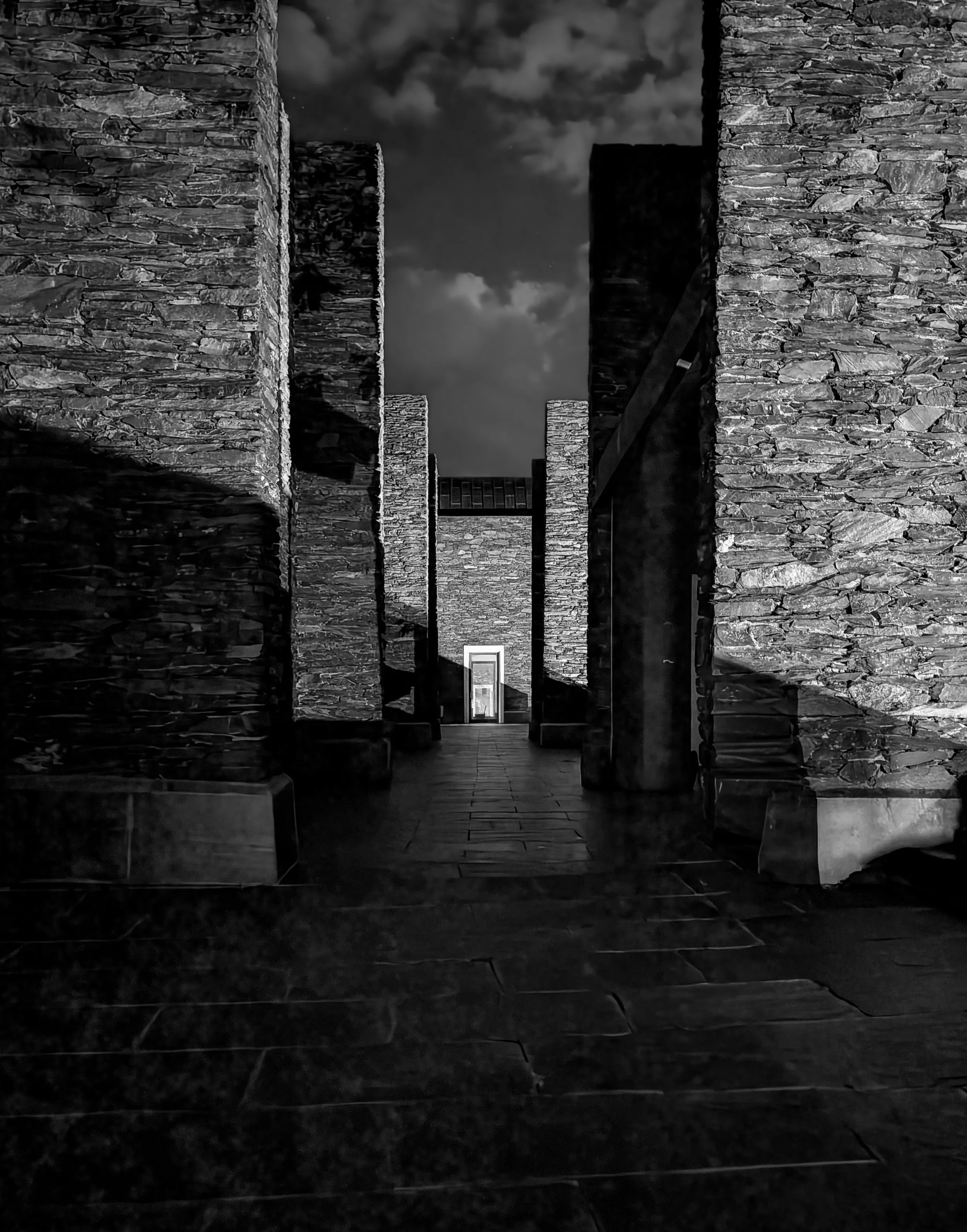 Black and white image of large stone walls and columns creating a heavy, grounded architectural space, with deep shadows and a distant opening.