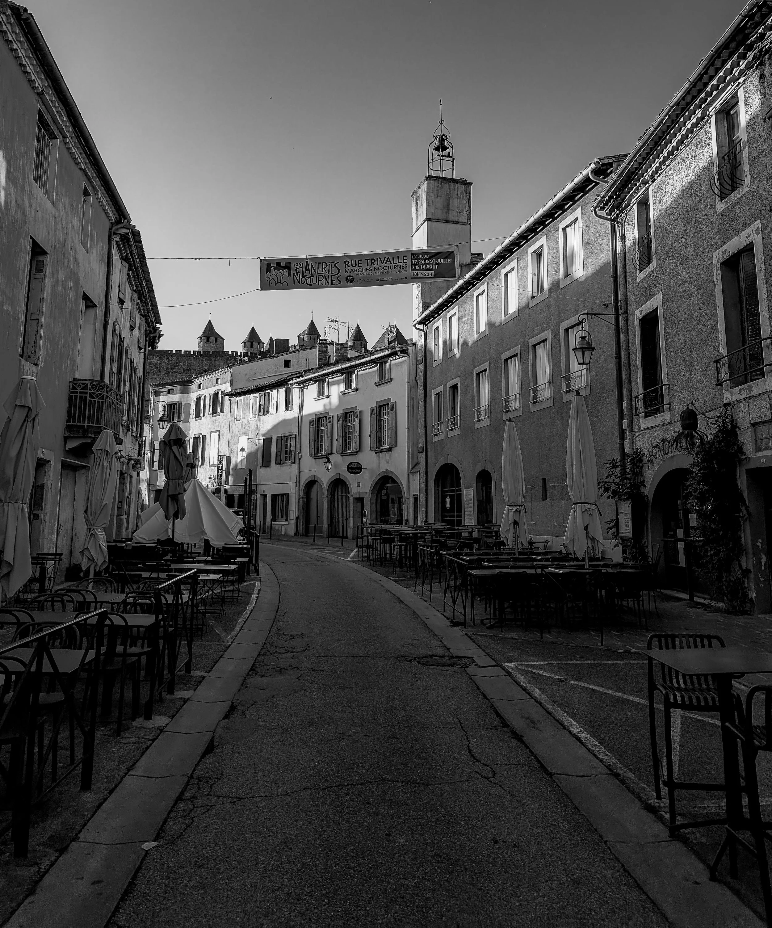 Black and white photograph of a quiet, curving street lined with buildings and empty café seating, guiding the eye through an open urban space.