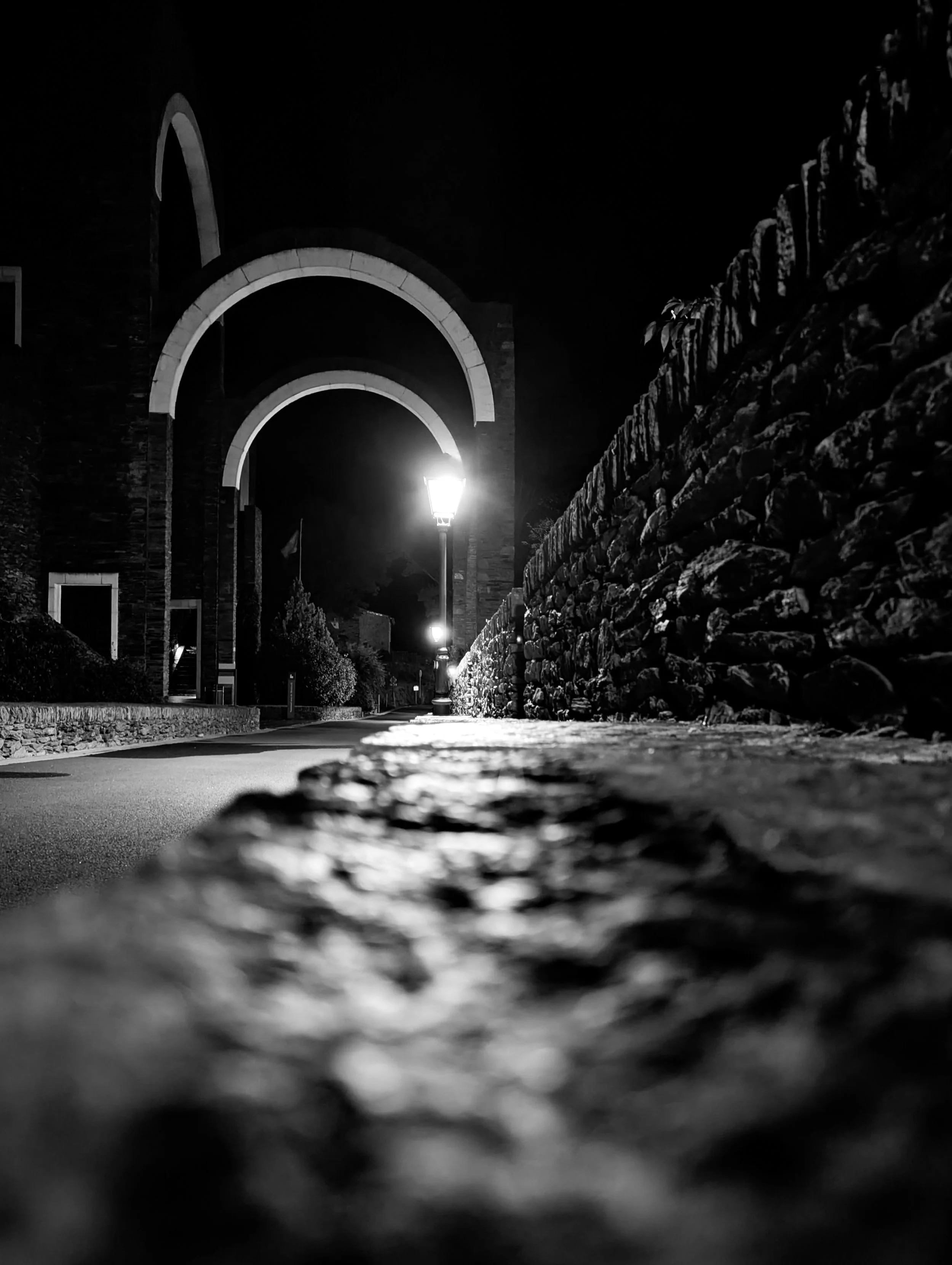 Night view of a cobblestone sidewalk illuminated by street lamps, with a stone wall on the right and an arched structure in the background.