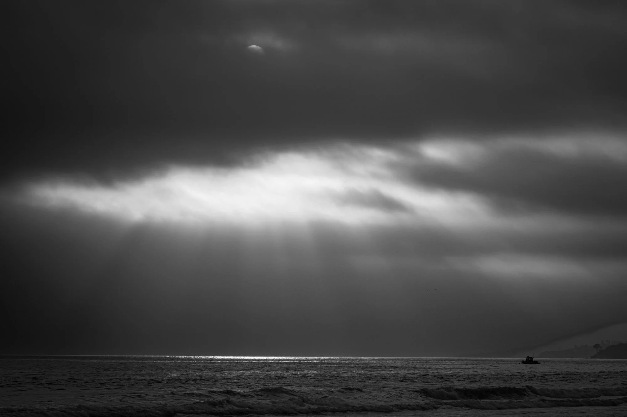 Black and white photo of the ocean with waves and a boat on the horizon under a cloudy sky, with the moon partially visible through clouds.