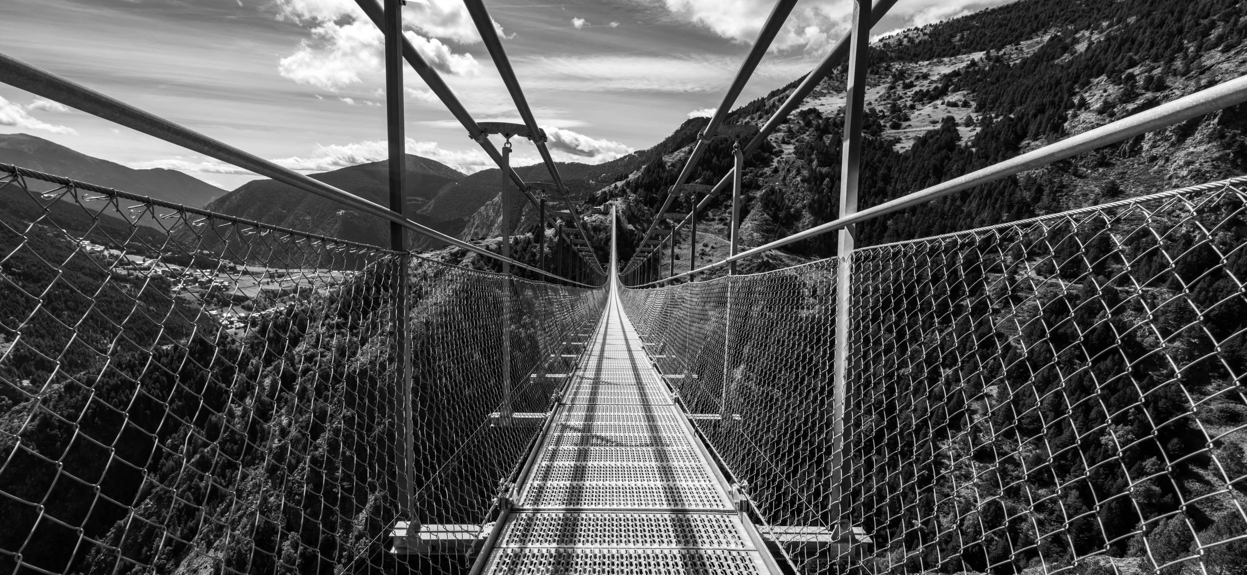A long suspension bridge made of metal with a mesh walkway and fencing on the sides, stretching over a deep valley with mountains and forested slopes in the background, under a partly cloudy sky.