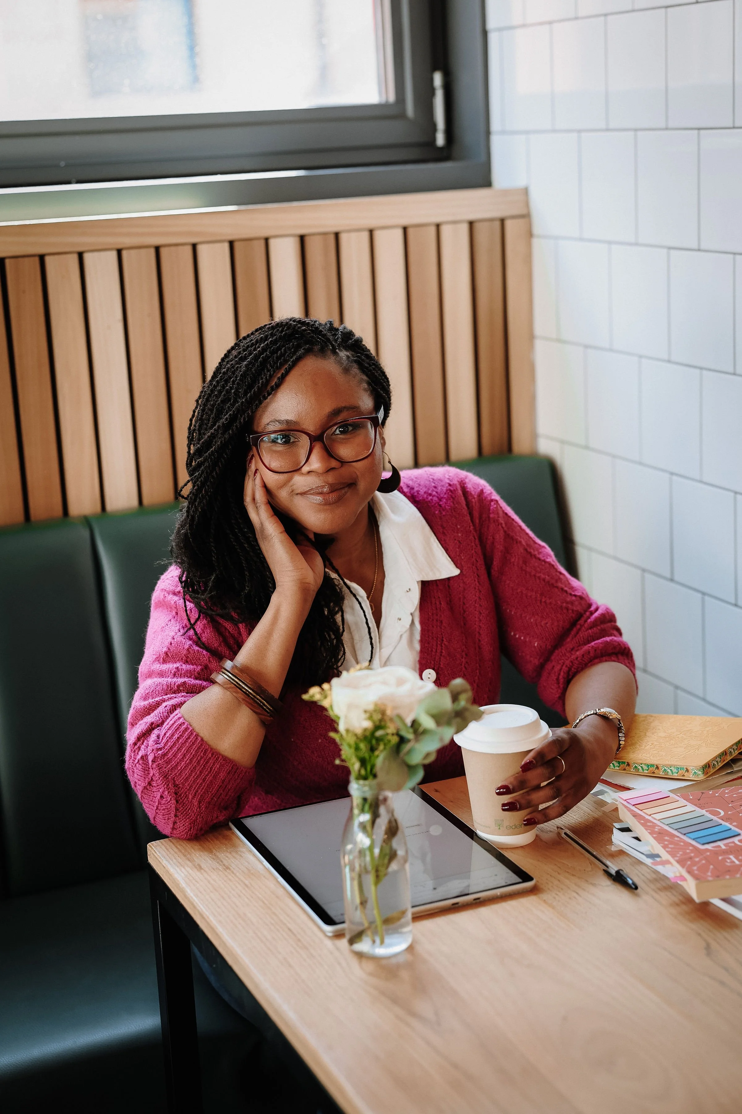 A wedding celebrant sits at a cafe with books of wedding poems, sticky notes, and a tablet. She's leaning on her hand and smiling at the camera, with her other hand around a coffee cup.