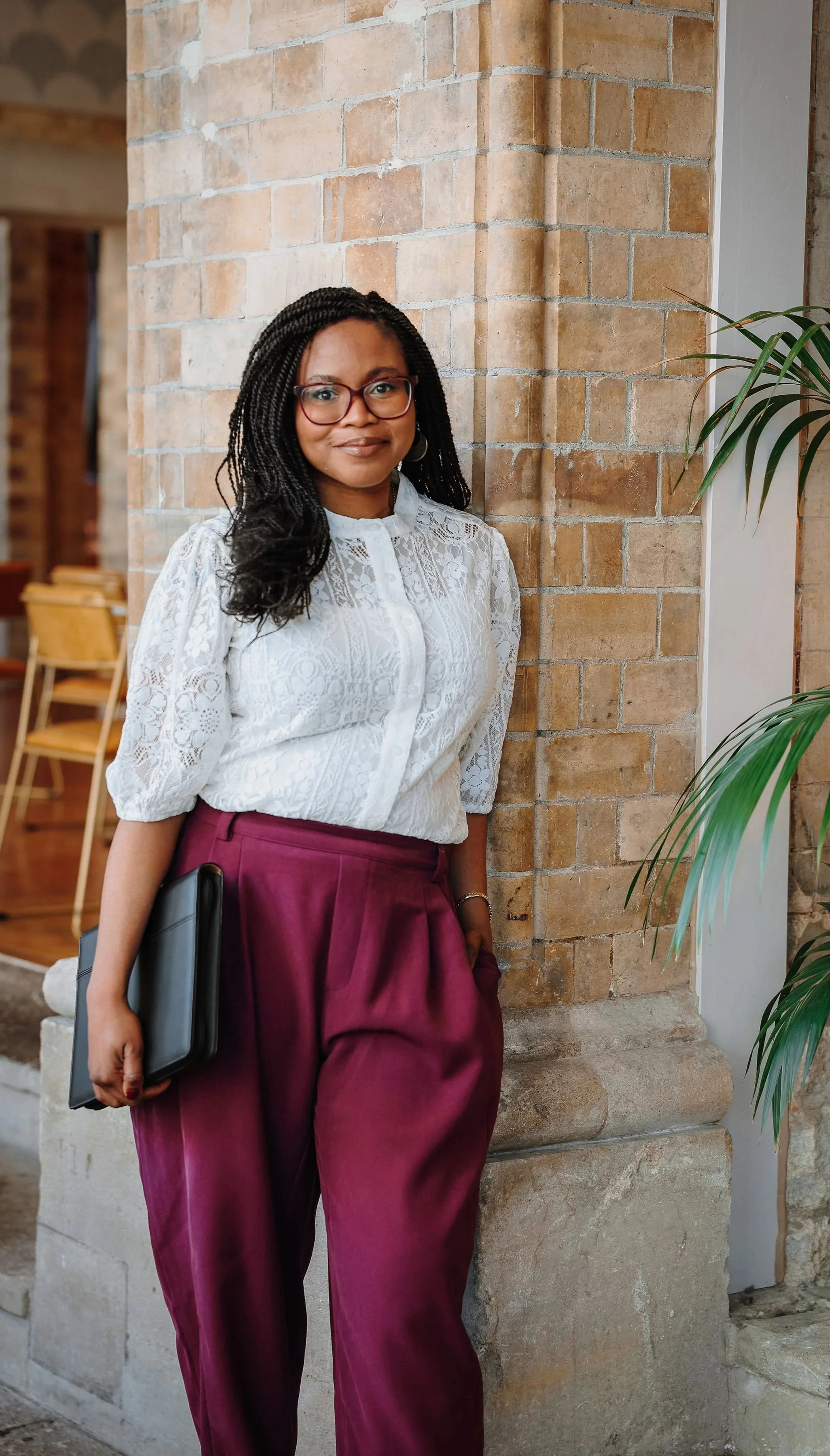 A wedding celebrant leans against a pillar, with a black folder in one hand. She is a Black woman with twists and red glasses.