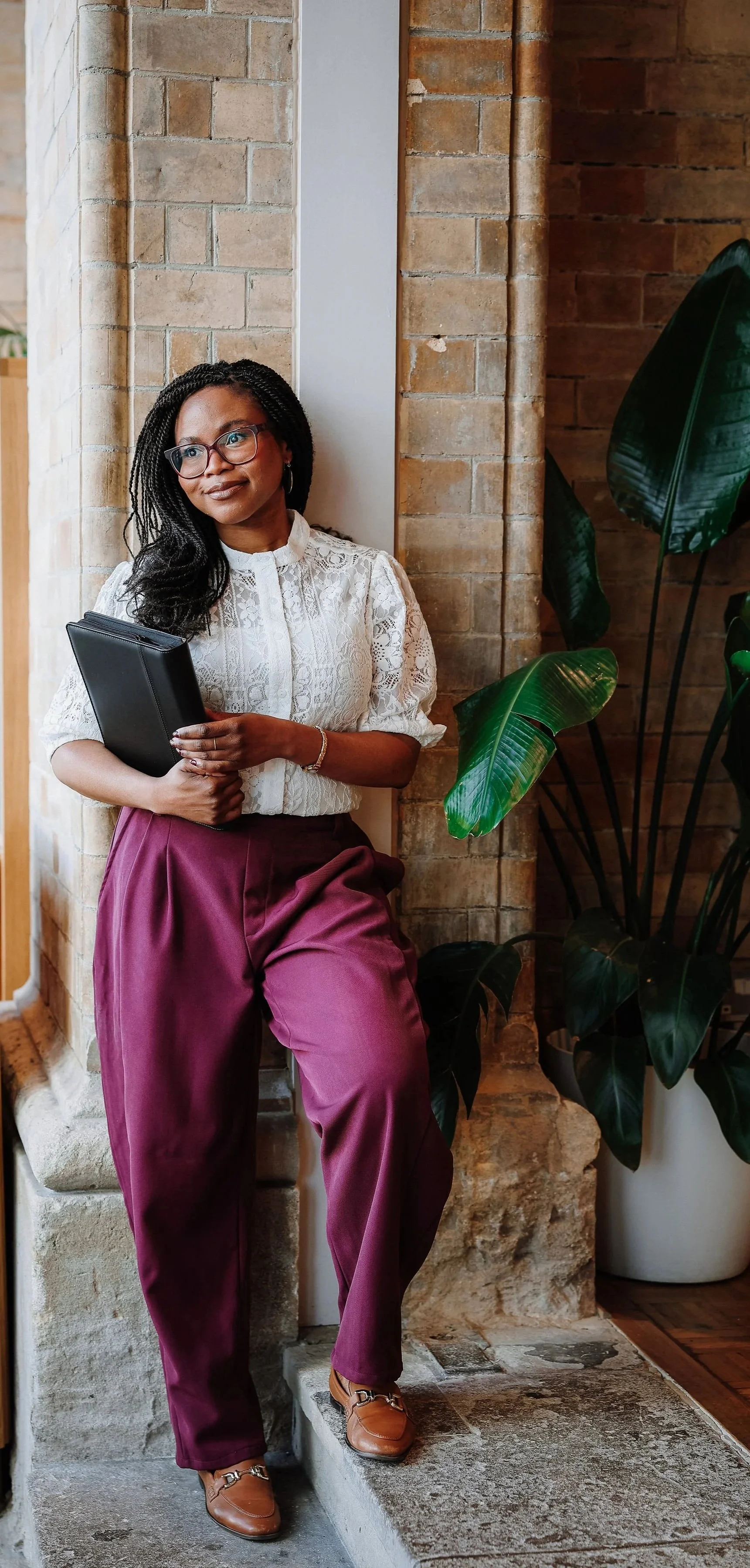 A wedding celebrant leans against a pillar and holds a black folder. She is a Black woman with glasses, and smiles softly while looking slightly away from the camera.