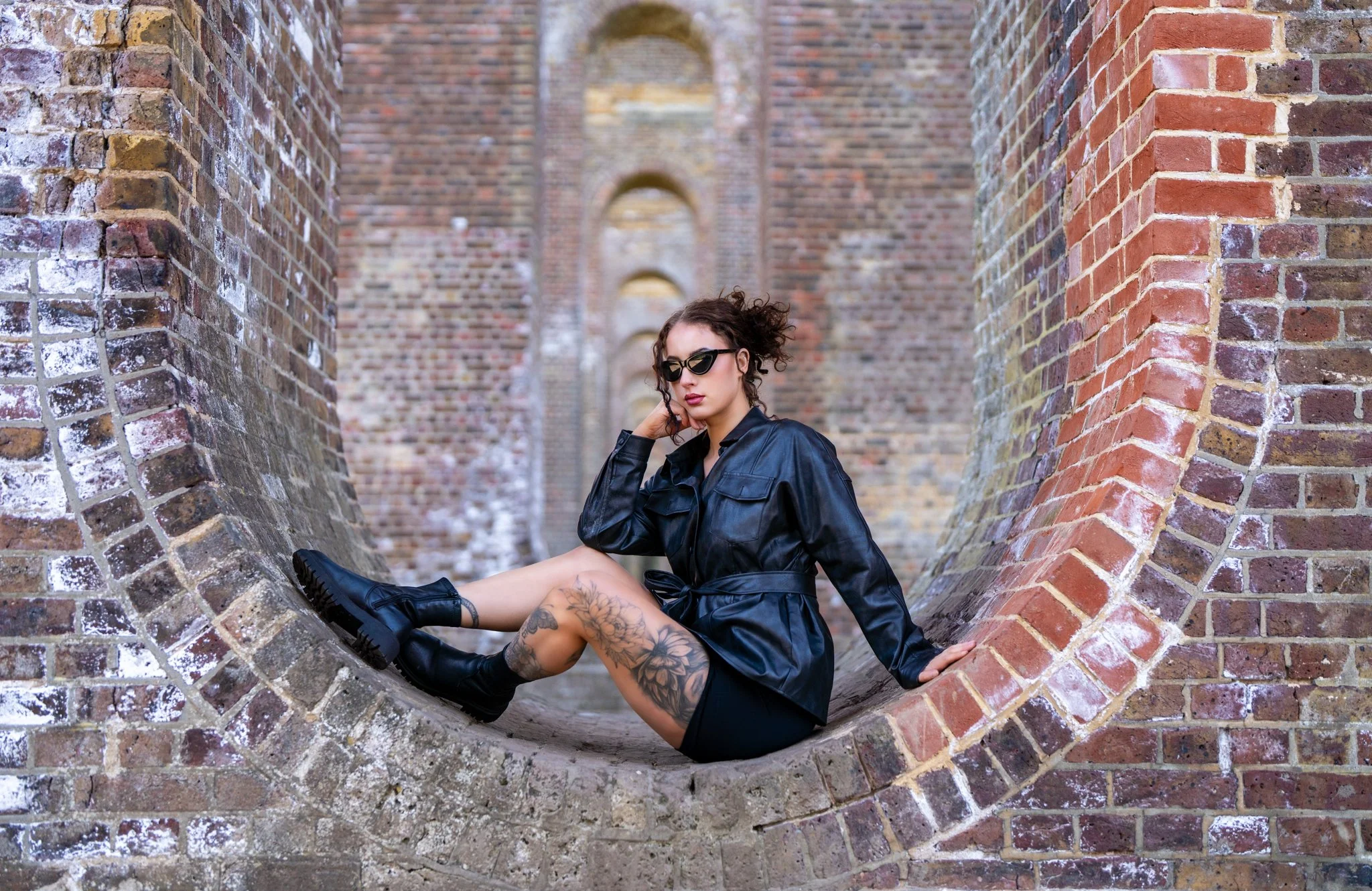 A woman with dark curly hair, wearing sunglasses and a black leather jacket, is sitting on a large brick structure with a circular opening, surrounded by brick walls, in an outdoor setting. Anne Wills Photography 