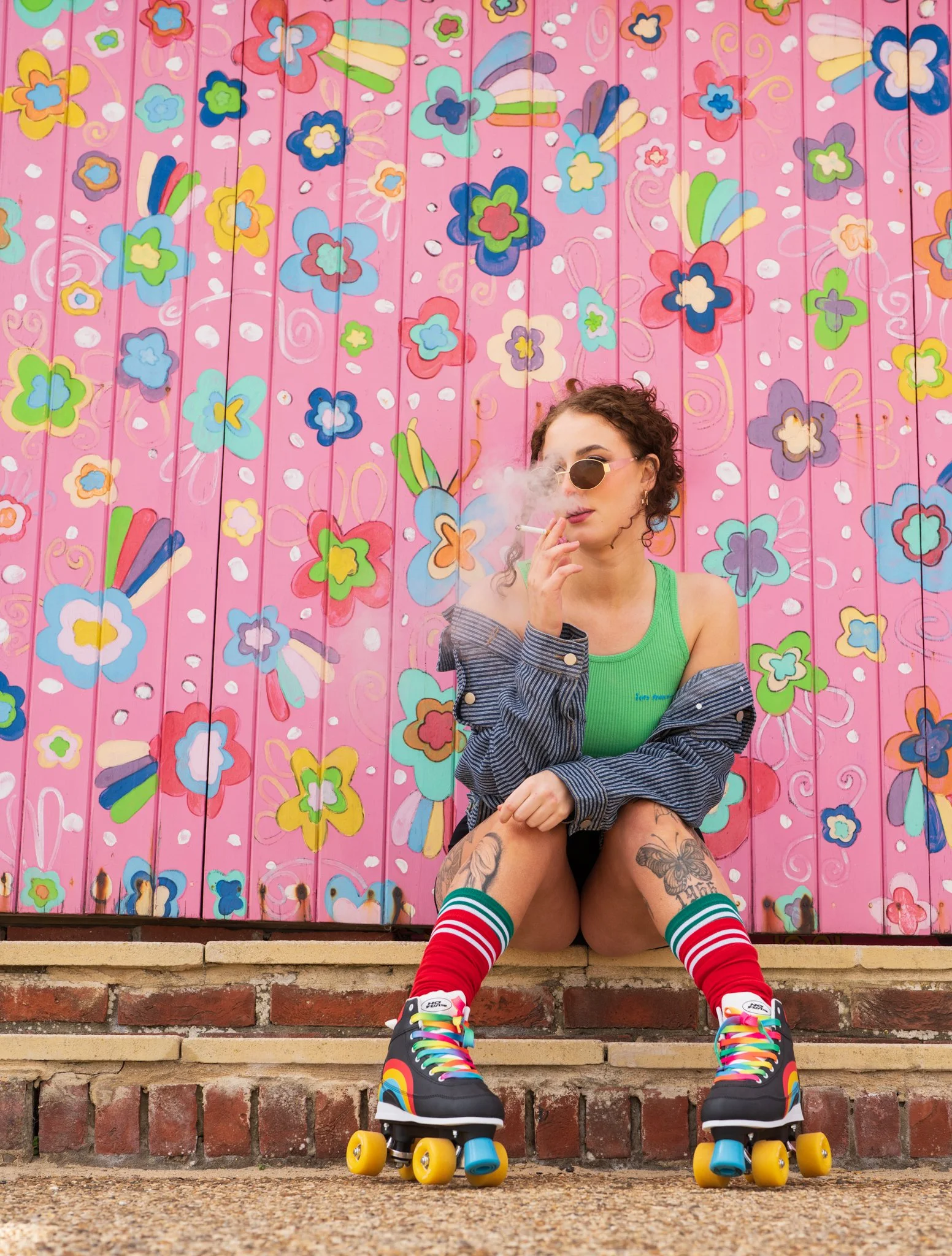 A woman wearing sunglasses, a green tank top, and striped socks is sitting on brick stairs in front of a colorful flower-themed mural, wearing roller skates with rainbow laces and yellow wheels while smoking a cigarette.
photographed at frinton, esse