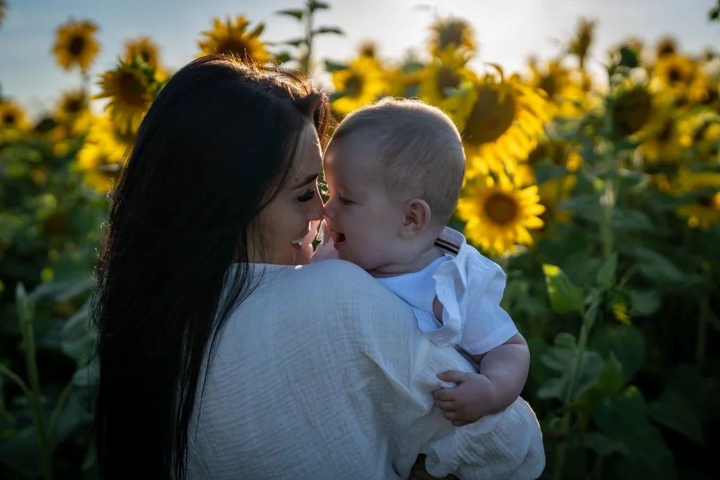 A woman holding a baby boy close to her face in a sunflower field during sunset.