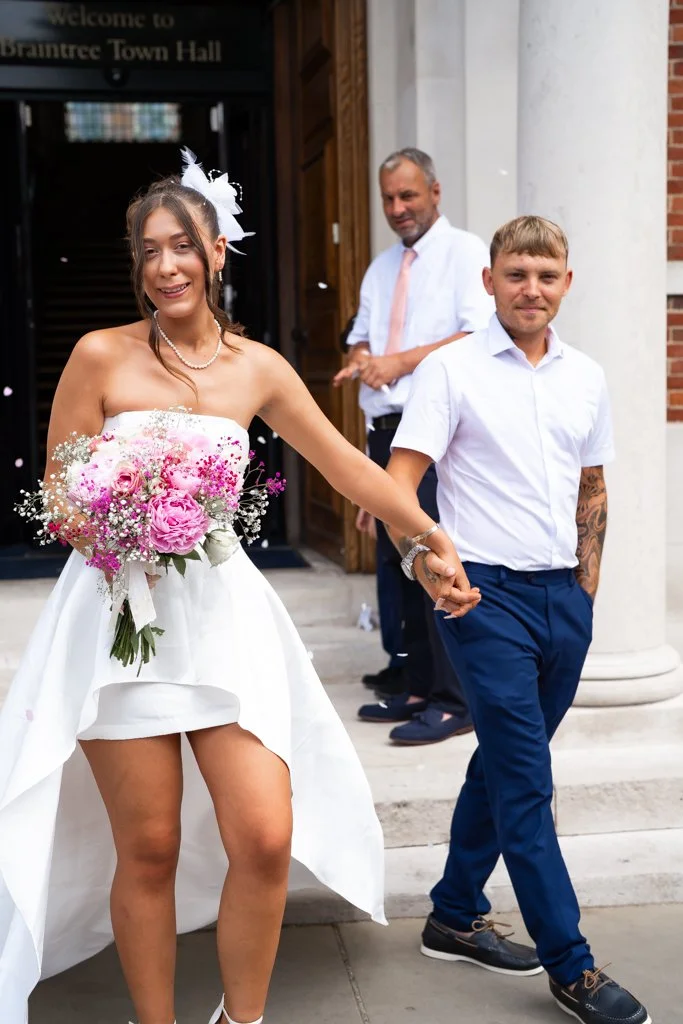 A bride in a white dress holding a pink and purple bouquet, smiling, holding hands with a groom in a white shirt and dark pants, standing outside Braintree Town Hall with an older man in a white shirt and light tie in the background. Anne Wills Photo