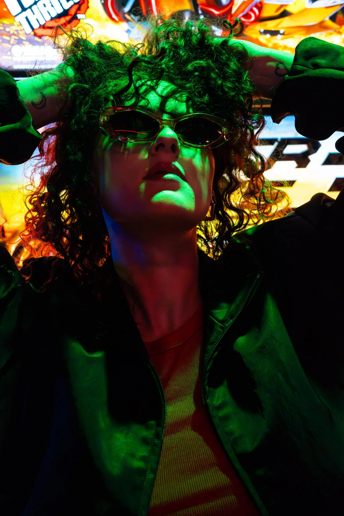 Person with curly hair wearing sunglasses, a green jacket, and a red shirt, with vibrant, colorful, neon lighting and signs in the background. photographed in Walton, essex in the arcades on the pier. Anne Wills Photography 
