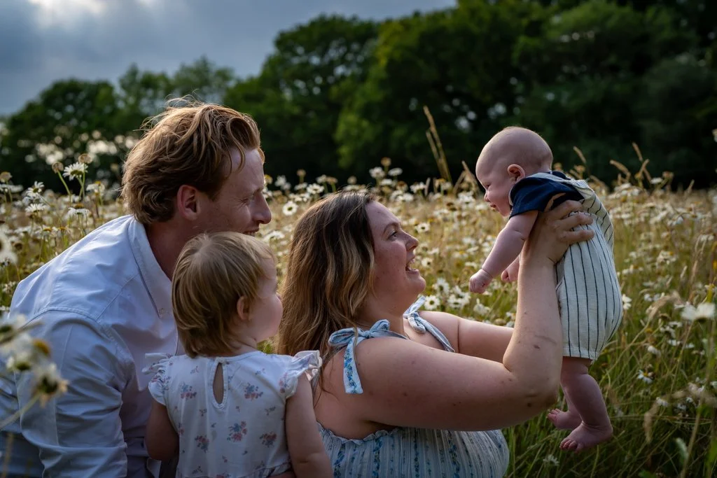 A family of four enjoying a joyful moment outdoors in a field of daisies, with trees in the background. The mother lifts a baby boy, while the father and a young girl watch and smile.