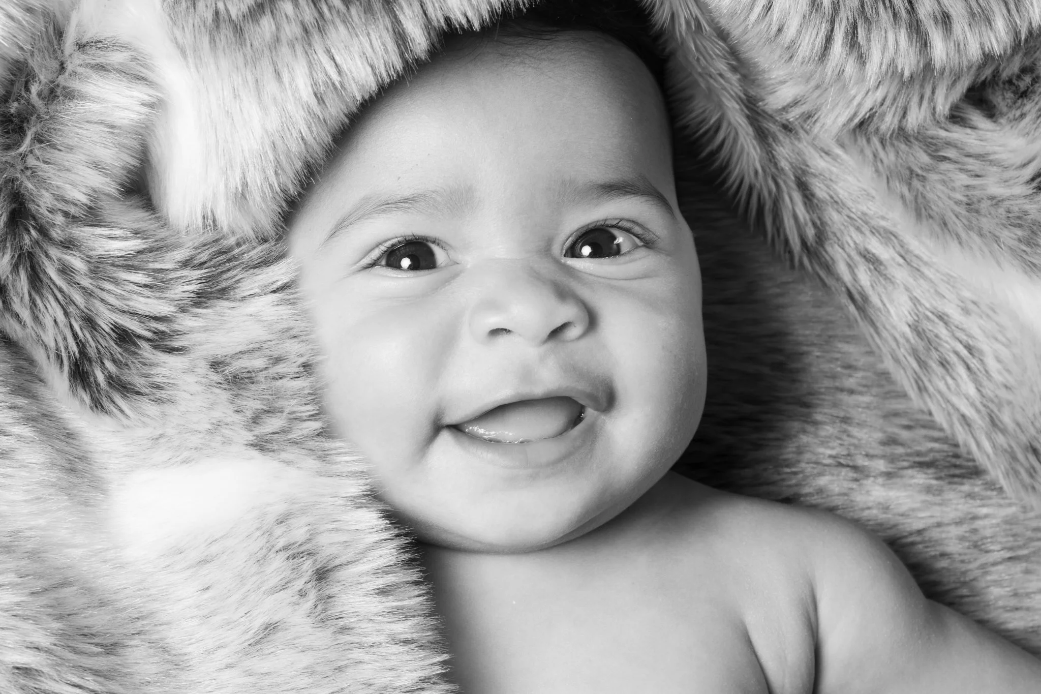 A smiling baby with wide eyes lying on a fur blanket.
