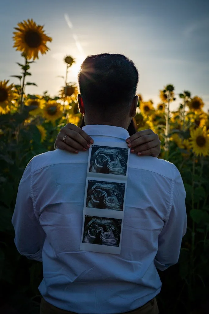 A man in a white shirt holding ultrasound photos behind his back while standing in a sunflower field at sunset.