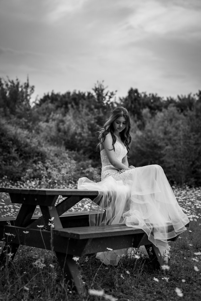 A black-and-white photo of a young woman in a wedding dress sitting on a wooden picnic table in a field with wildflowers, surrounded by trees, under a partly cloudy sky.