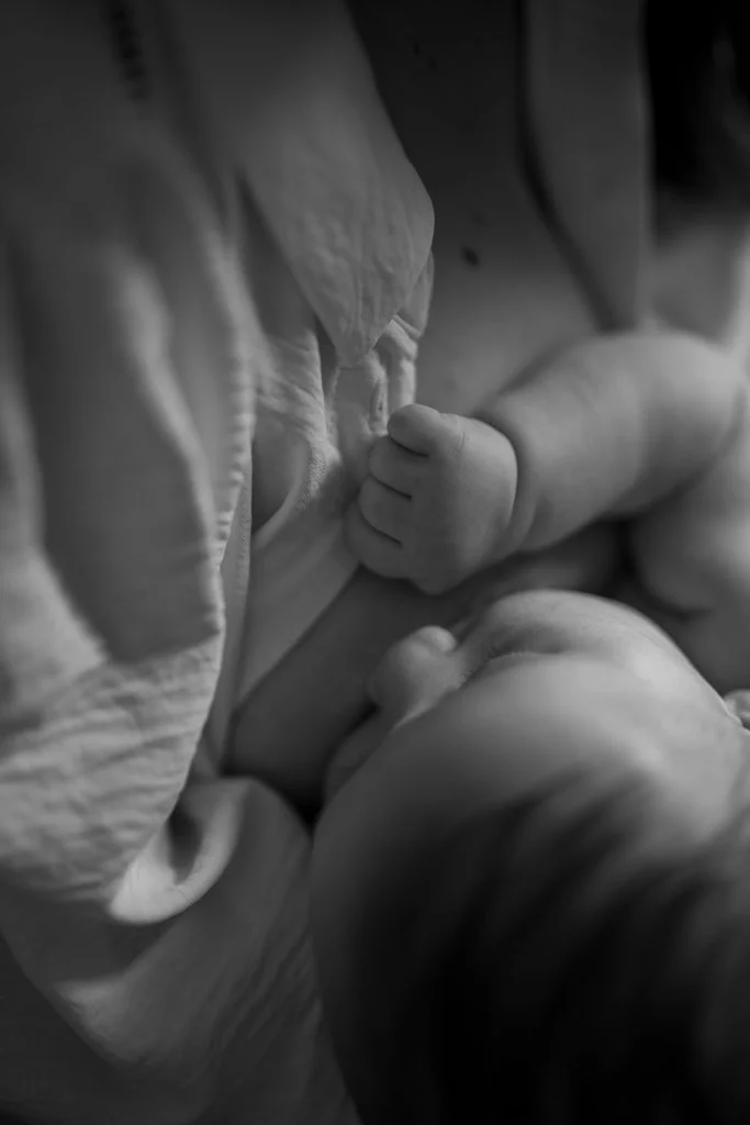 Close-up black-and-white photo of a baby nursing from an adult, showing the baby's hand and the adult's chest.
