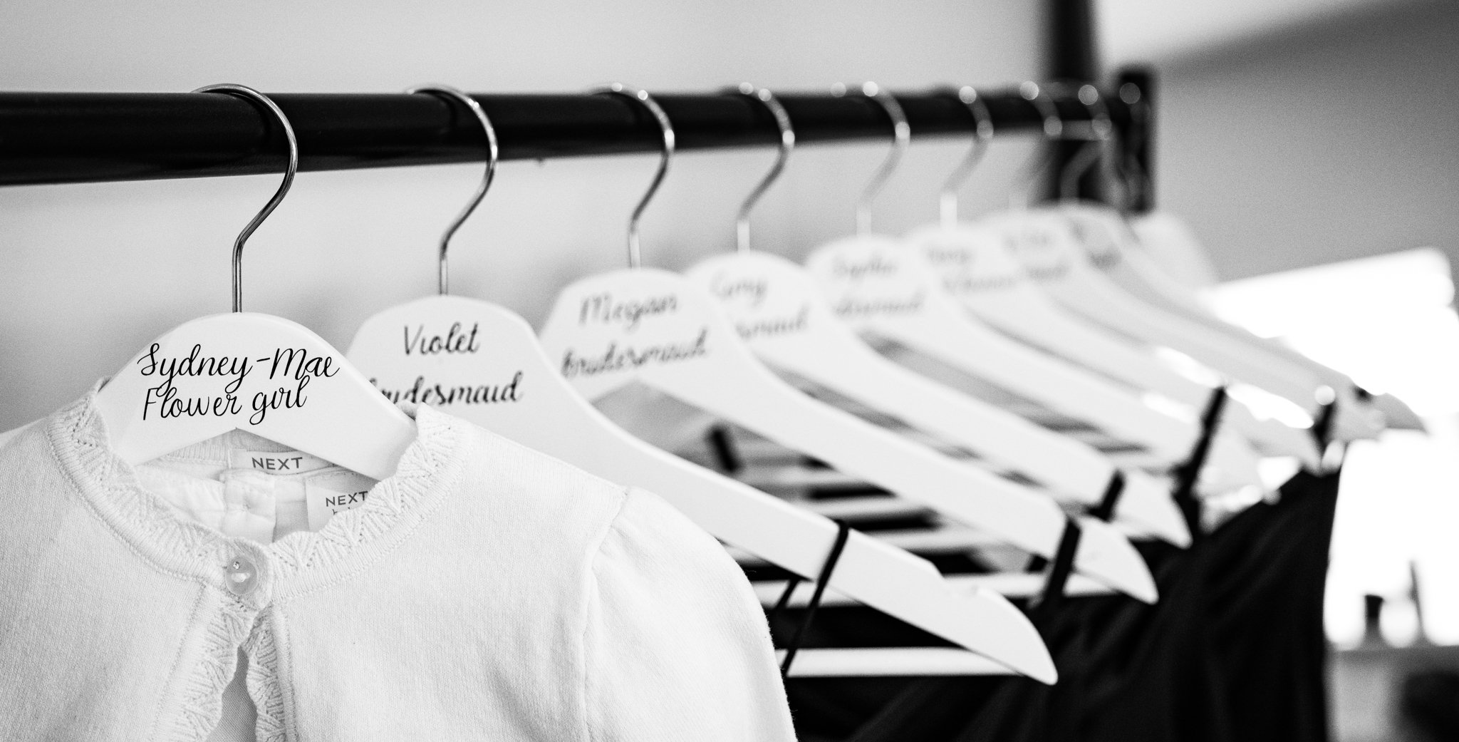 Black and white photograph of a row of white clothes hangers on a black clothing rack, with a white child's sweater hanging on the closest hanger. The sweater has a label that reads "NEXT" and a ruffled collar with a button. The hangers are labeled w