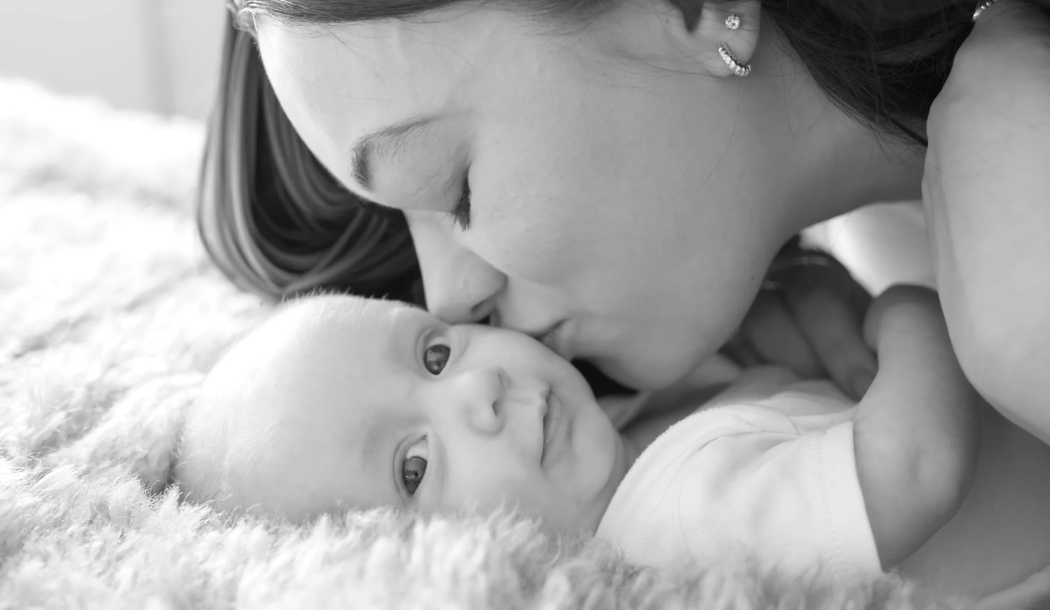 A black and white photo of a woman gently kissing a baby's forehead as they lie on a soft blanket, both with light skin, in a tender moment.