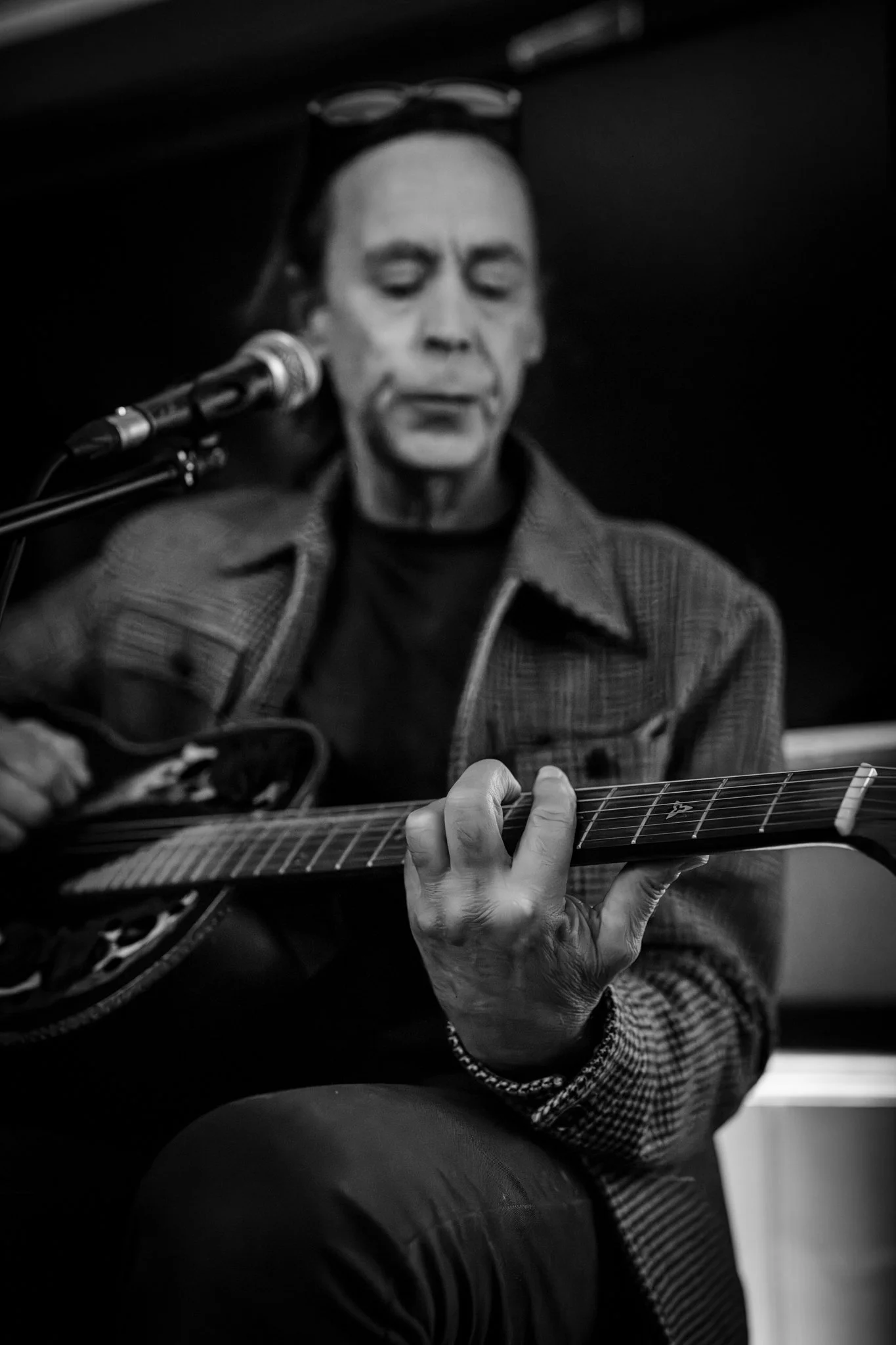A person playing an acoustic guitar, wearing a checked jacket, with glasses resting on their head, in a black and white setting. Anne Wills Photography. Braintree, Essex