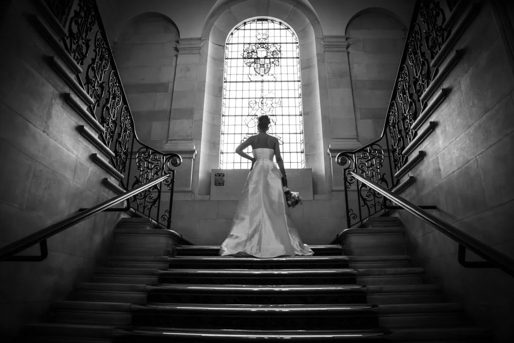 A woman in a wedding dress holding a bouquet standing on a grand staircase inside a church, facing a tall stained glass window. Anne Wills Photography. Braintree town hall, essex