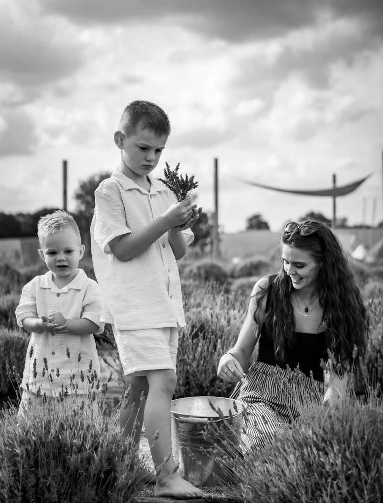 A woman with long hair and sunglasses on her head is kneeling in a lavender field, smiling, while two young boys are standing nearby, one holding a small branch and the other looking at the camera, with a bucket and lavender plants around them.