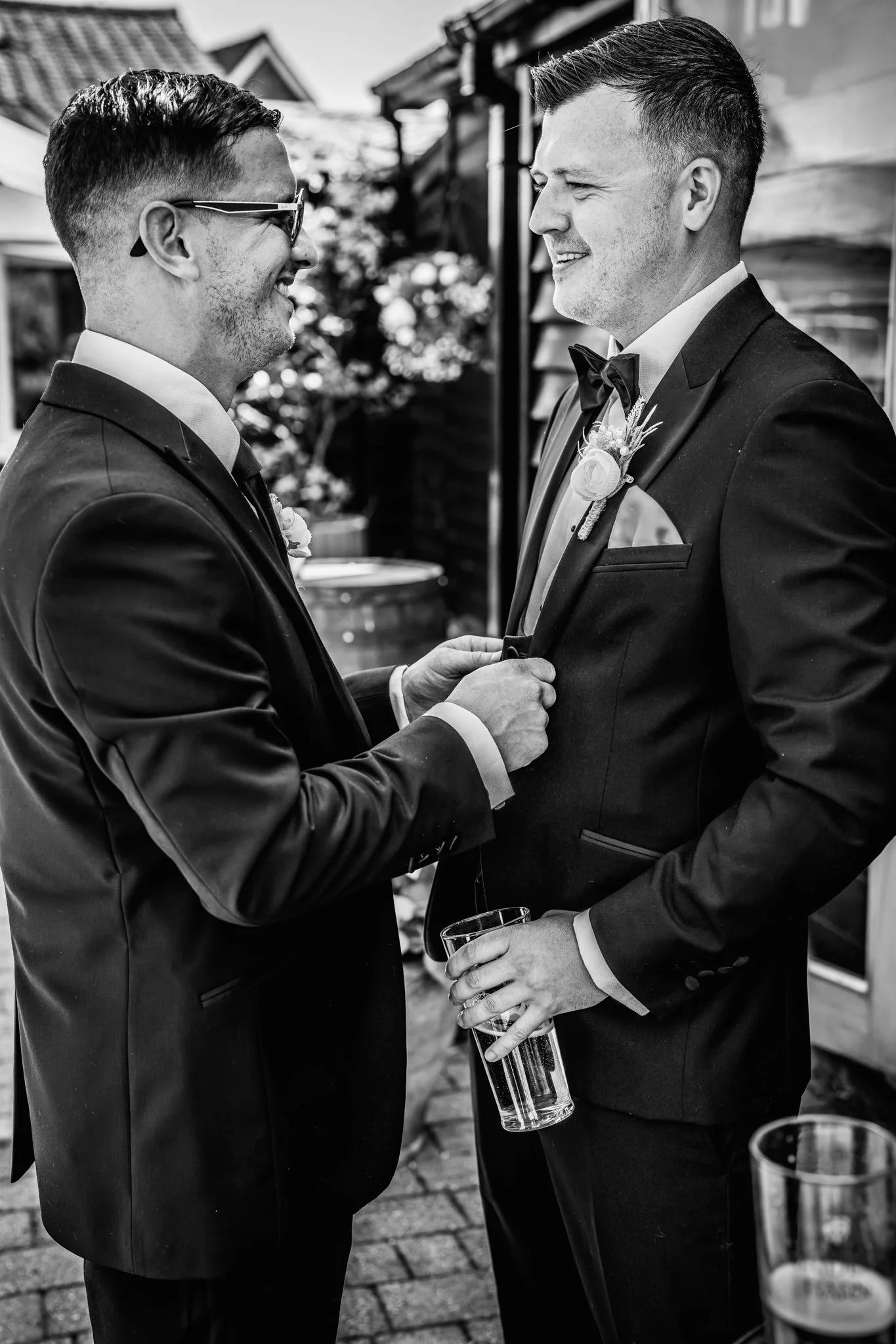 Two men in tuxedos face each other, smiling, in a close interaction at an outdoor event, with drinks in their hands. Anne Wills Photography Maidens barn Essex