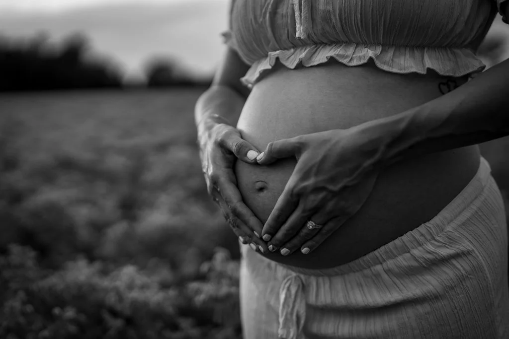 A pregnant woman is holding her belly with both hands, with a partner's hands gently framing her belly in an outdoor field setting.