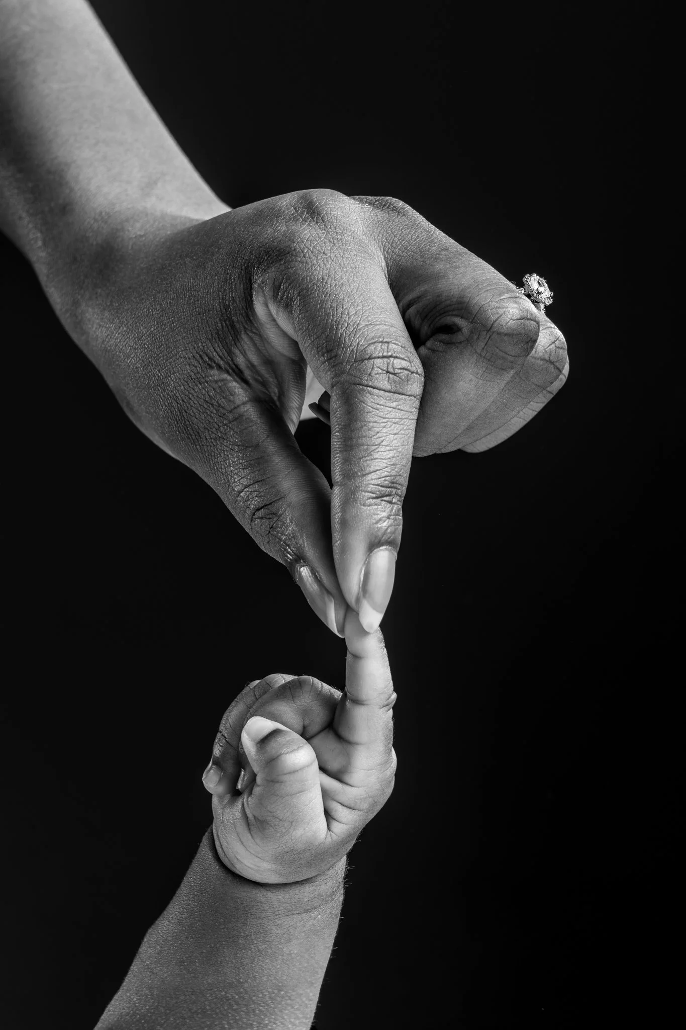 A black and white photograph of two hands; one adult's hand with a ring, one child's hand,  intertwined and touching in a delicate manner against a black background.