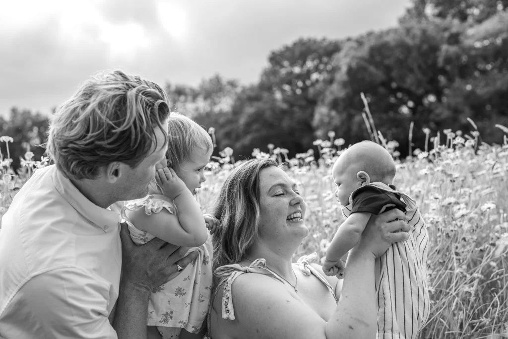 A family outdoors in a field of flowers, with two adults, a young girl, and a baby, all smiling and interacting happily. Anne Wills Photography. Braintree, Essex