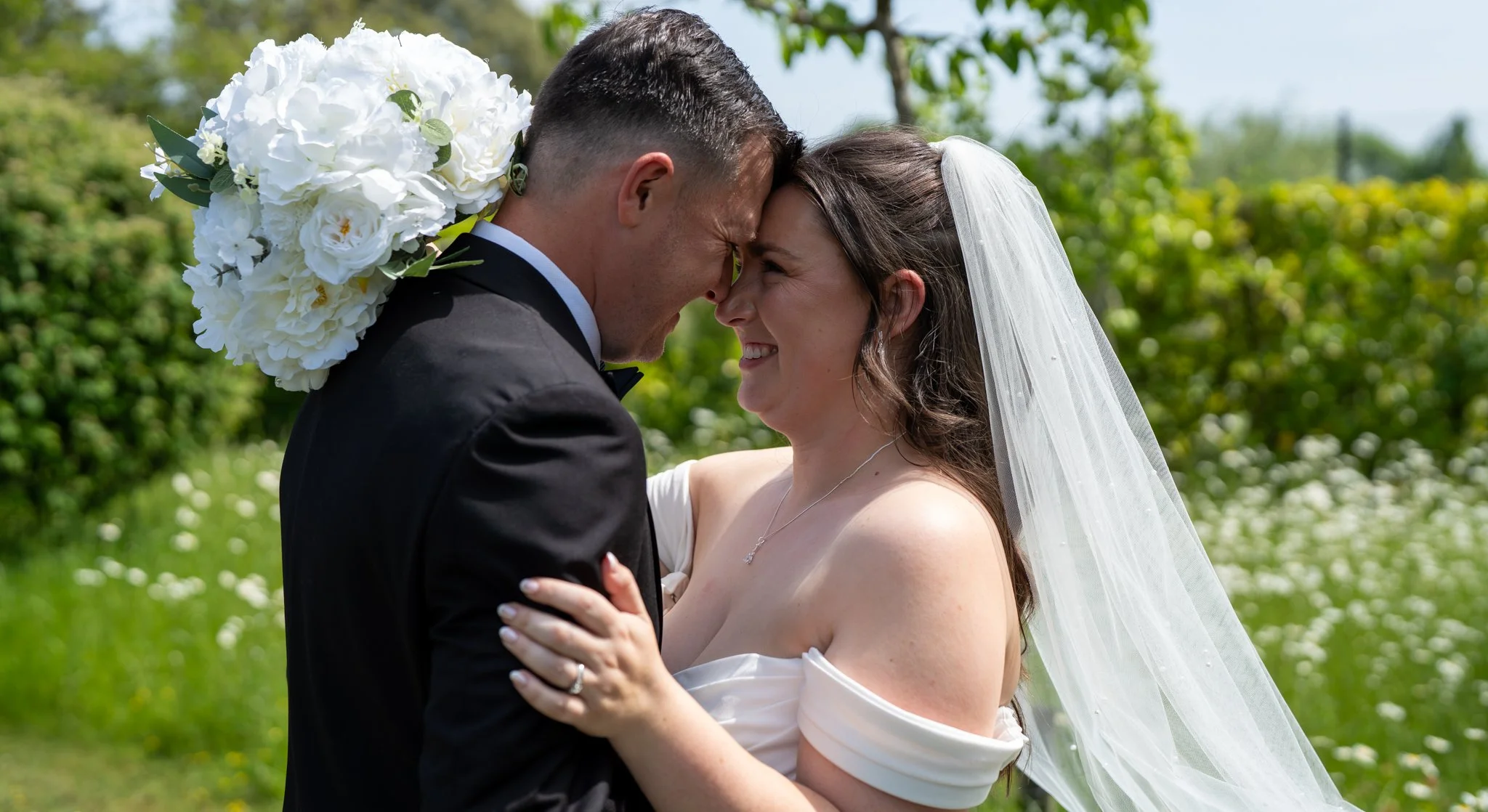 A bride and groom are smiling and touching foreheads outdoors on their wedding day, with a bouquet of white flowers on the groom's shoulder. Anne Wills Photography Maidens barn Essex
