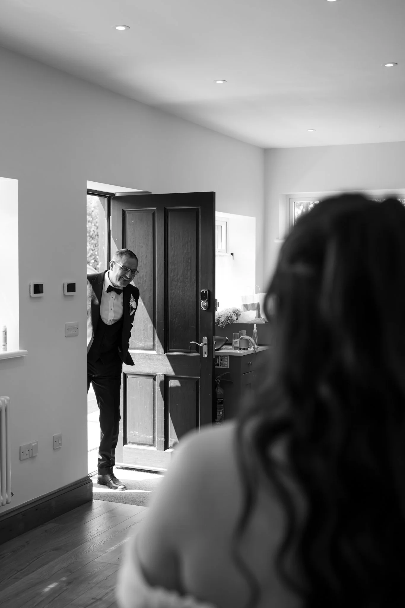 A man in a tuxedo, possibly at a wedding or formal event, is smiling and standing just inside a door in a room with wooden floors. There are people in the foreground with blurred heads, and sunlight is streaming in through a window. Anne Wills Photog