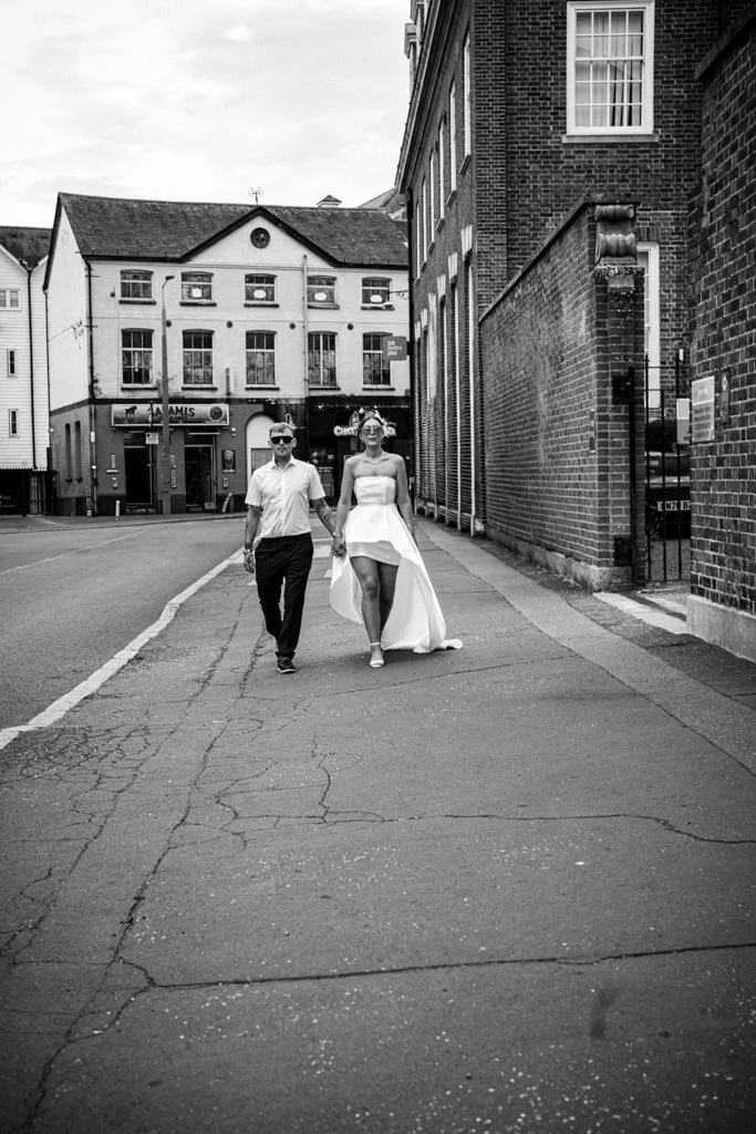 A couple holding hands walking on a city sidewalk, woman in a white dress and man in a white shirt and dark pants, black and white photo. Anne Wills Photography. Braintree town hall, Essex