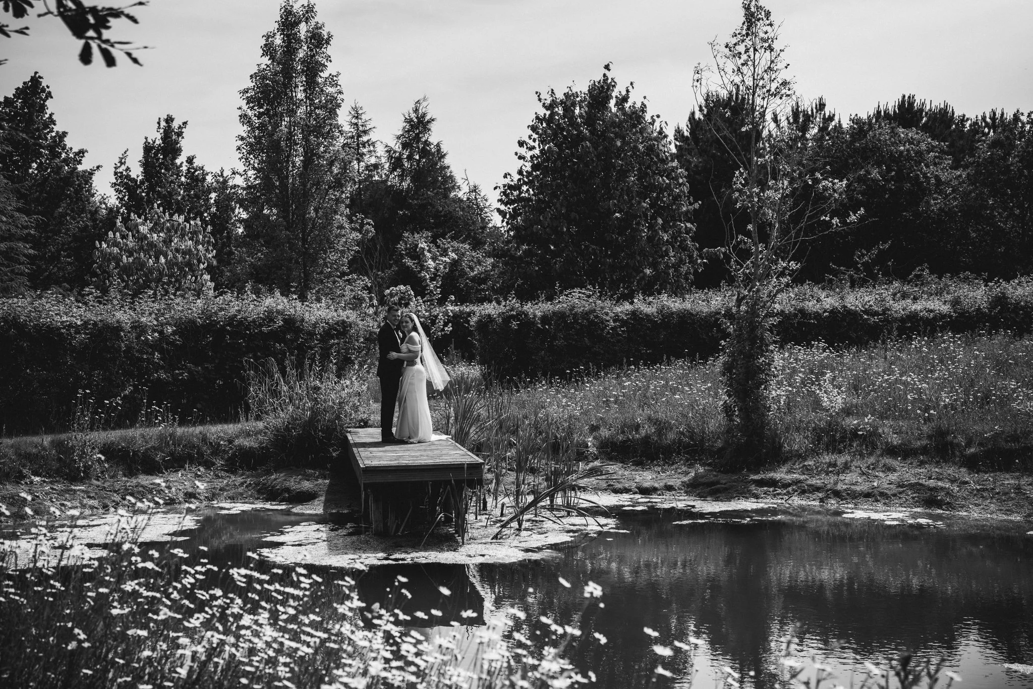 A black and white wedding photo of a couple standing on a small wooden bridge in a natural outdoor setting, surrounded by trees and a pond. Anne Wills Photography Maidens barn Essex