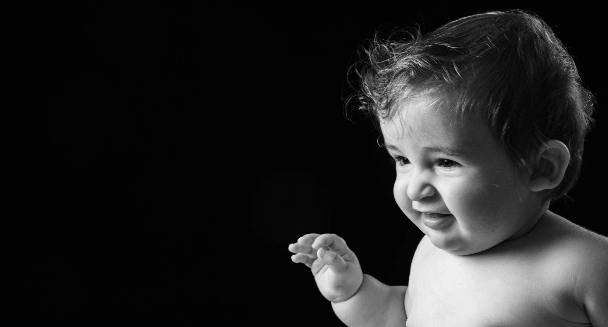 A black and white photo of a smiling young child with curly hair, looking to the right, with hands clasped. Anne Wills Photography 