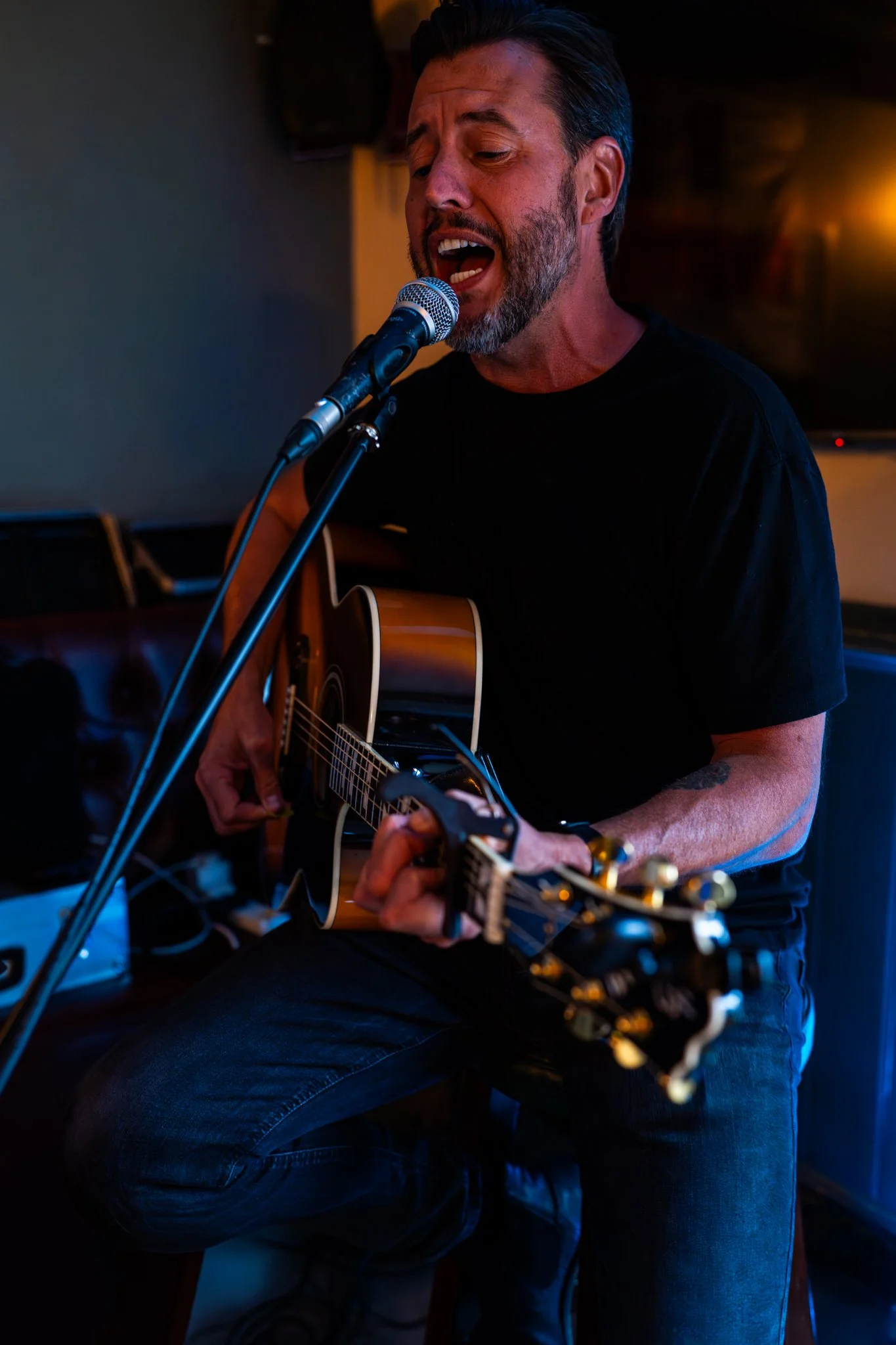 A man singing passionately into a microphone while playing an acoustic guitar, with a warm orange light illuminating the background. Anne Wills Photography. Braintree, Essex