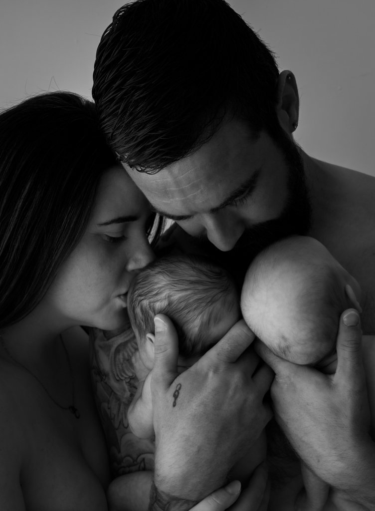 A black and white photograph of a family of three holding their newborn baby. The mother and father are tenderly kissing and supporting the newborn's head and body.