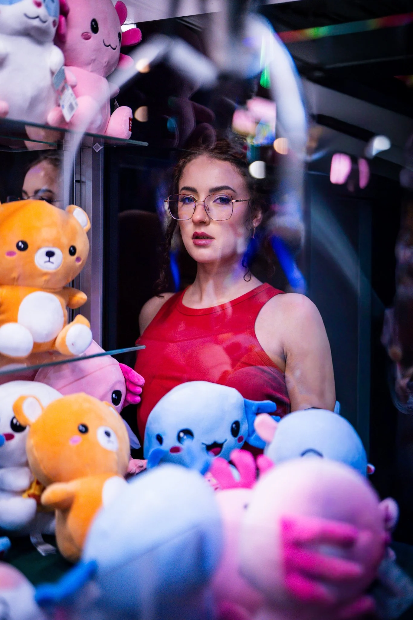 A young woman with glasses and curly hair wearing a red dress looking through a glass display of colorful plush toys in a claw machine. The plush toys include a pink character, a blue character, and a brown bear, with some toys reflected on the glass