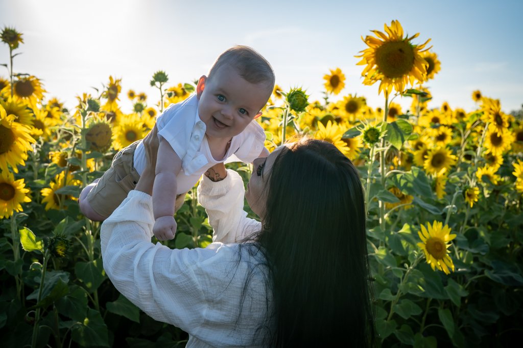 A woman lifting a smiling baby in a sunflower field during sunset.