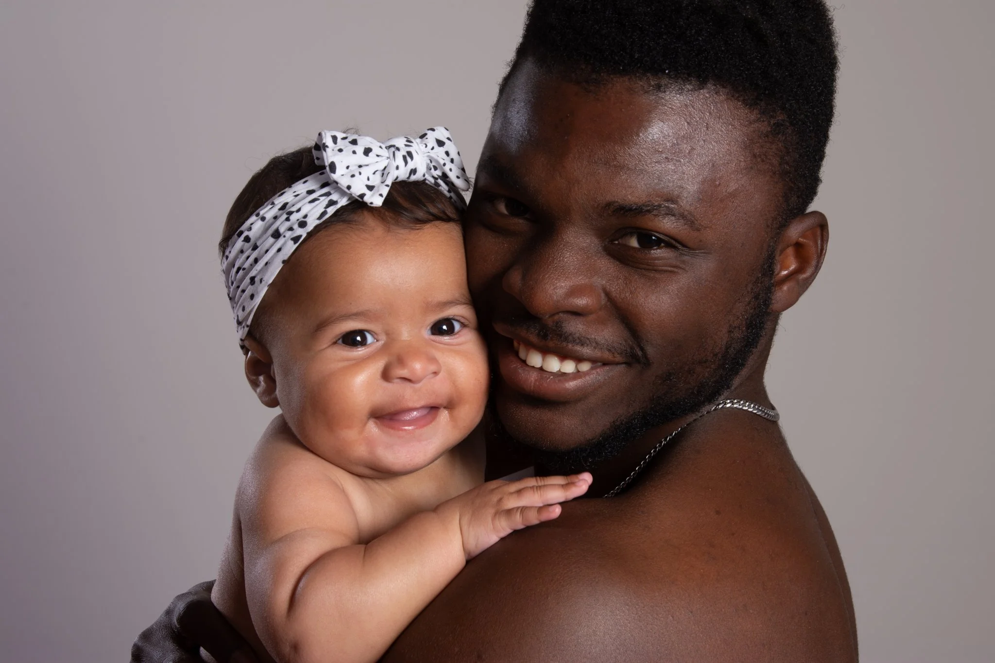 A smiling Black man holding a happy infant girl with a polka dot headband, both close together against a plain gray background. Anne Wills Photography 