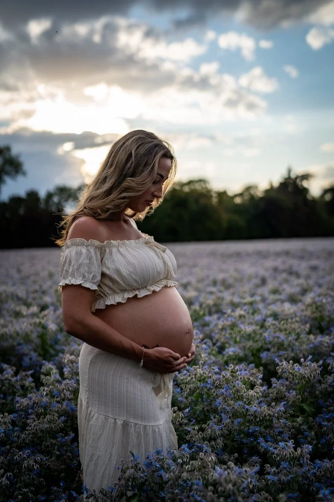 Pregnant woman standing in a field of purple flowers during sunset, gazing down at her belly.