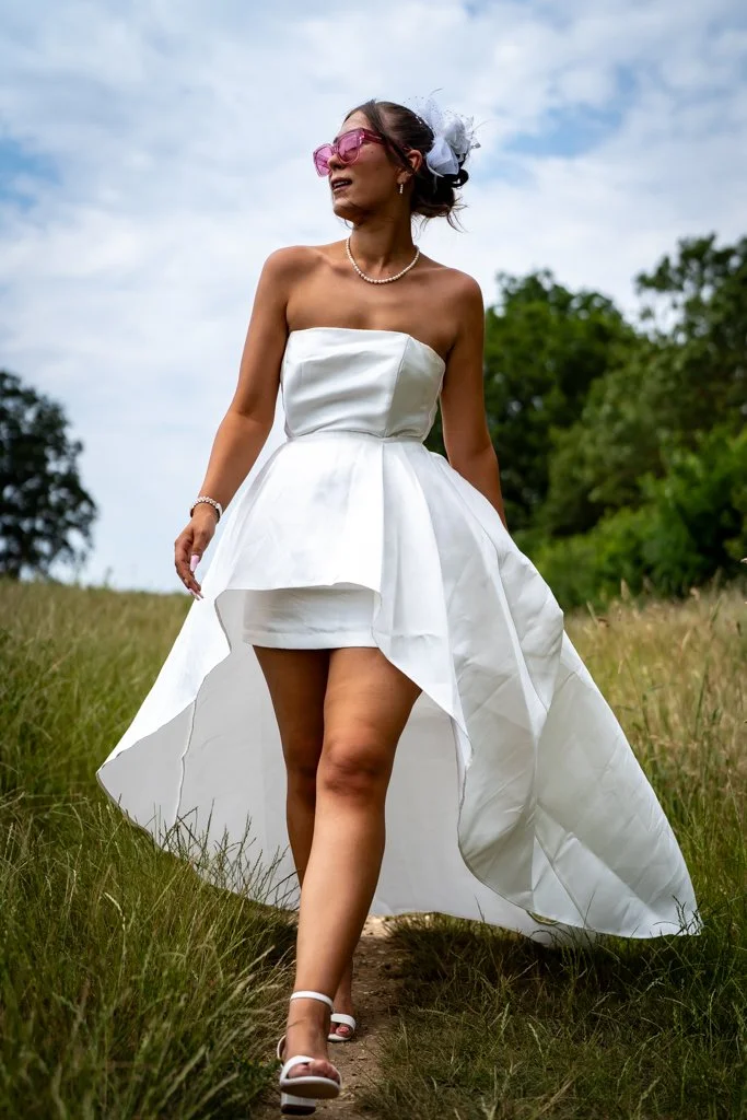 A woman in a strapless white wedding dress walking outdoors on a grassy trail, wearing pink sunglasses, a pearl necklace, a white hair accessory, and open-toe white sandals. Anne Wills Photography. Braintree, essex