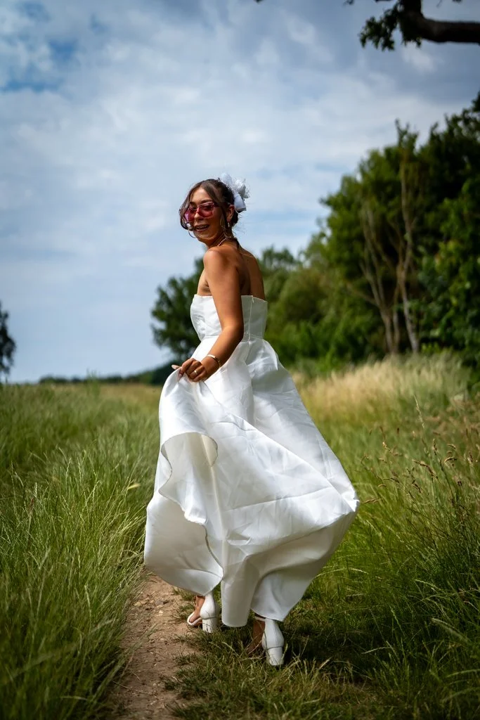 Woman in a white wedding dress standing on a grassy path in a natural outdoor setting, smiling and wearing sunglasses. Anne Wills Photography. Braintree town hall, Essex