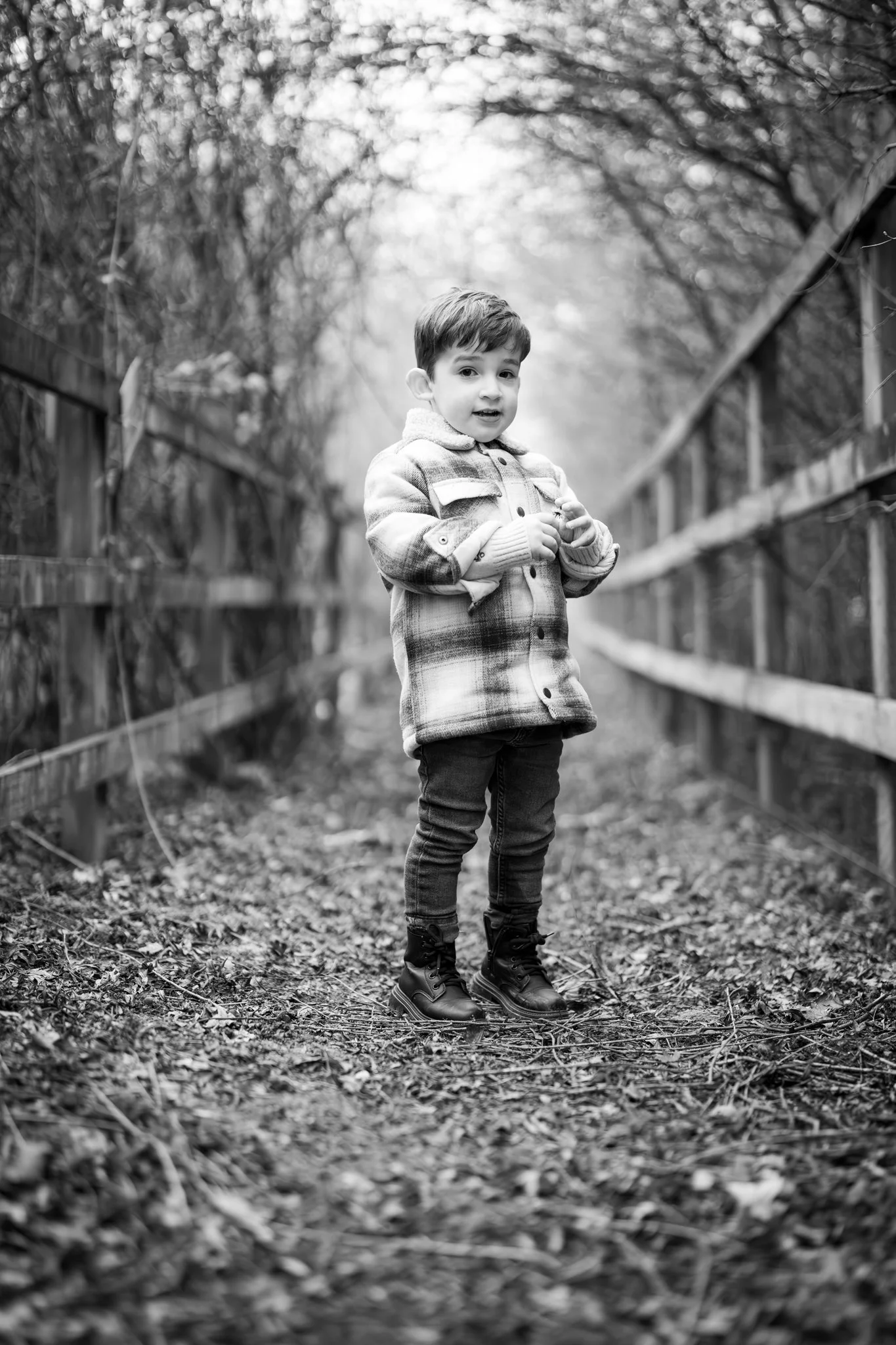 A young boy standing on a leaf-covered path between wooded fences, wearing a jacket and boots, captured in black and white. photographed in Dunmow, Essex. Anne Wills Photography 