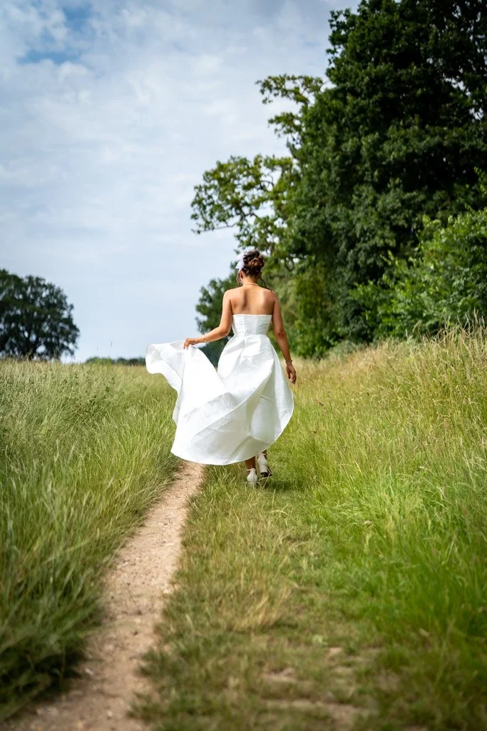 Woman in a white dress walking down a grassy dirt path surrounded by trees on a partly cloudy day. Anne Wills Photography. Braintree town hall, Essex